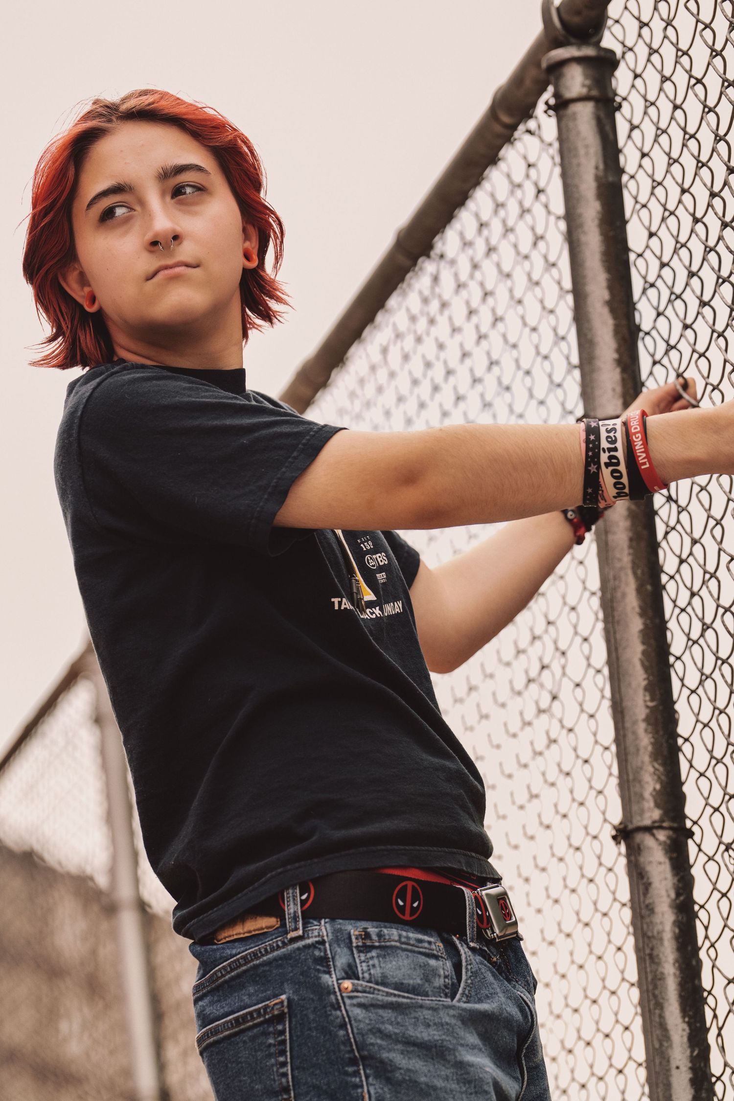 A person with red hair in a black t-shirt and jeans poses against a chain link fence outdoors.
