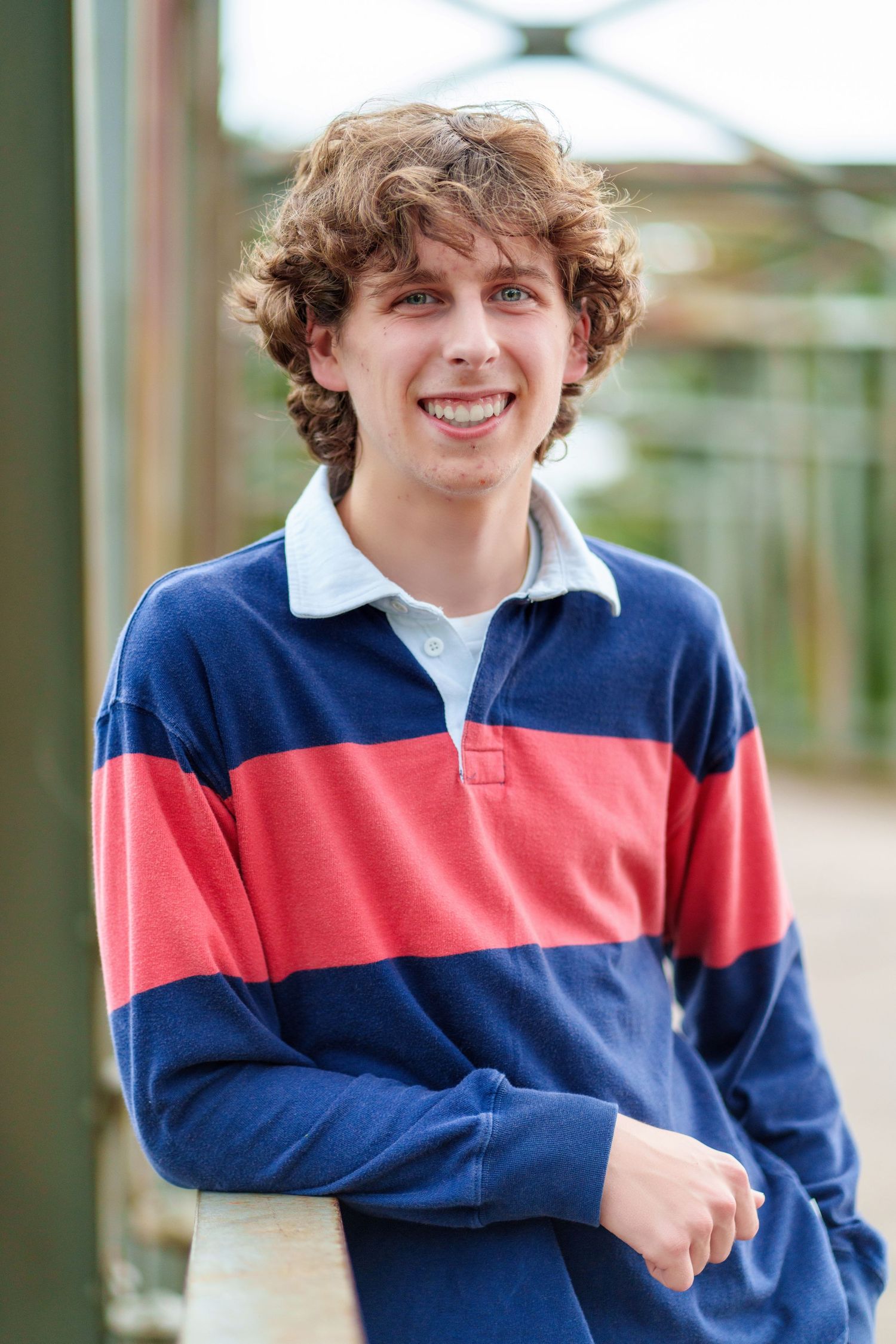Smiling teenage boy with curly hair wearing a blue and red striped rugby polo shirt, leaning on a railing outdoors.