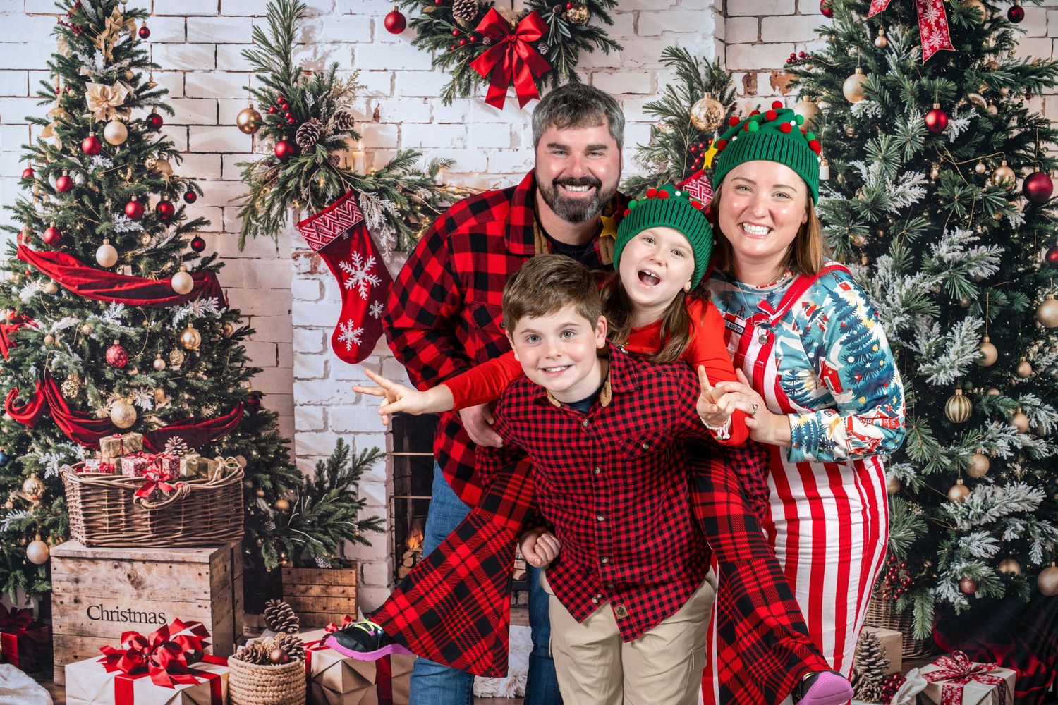 Family poses in coordinated red and black buffalo plaid outfits against a decorated Christmas backdrop with brick wall.