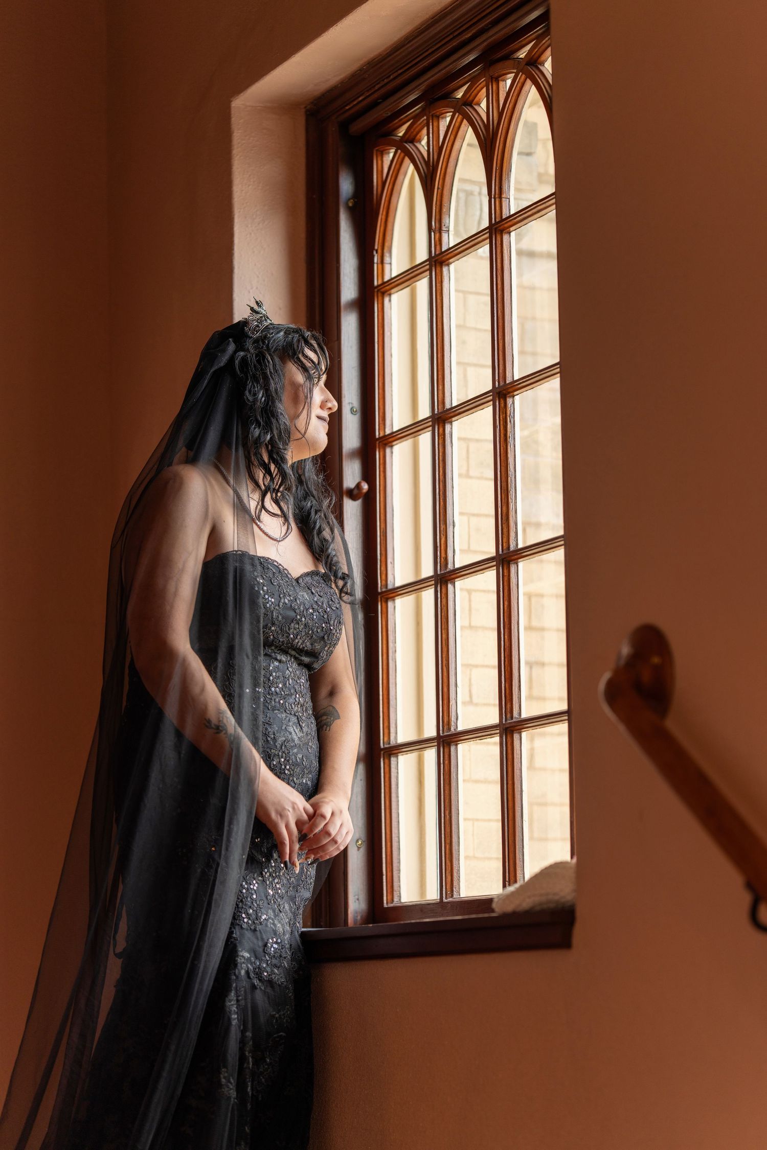 A person in a black veil poses dramatically by arched windows in a warm-lit room with gothic architectural elements.