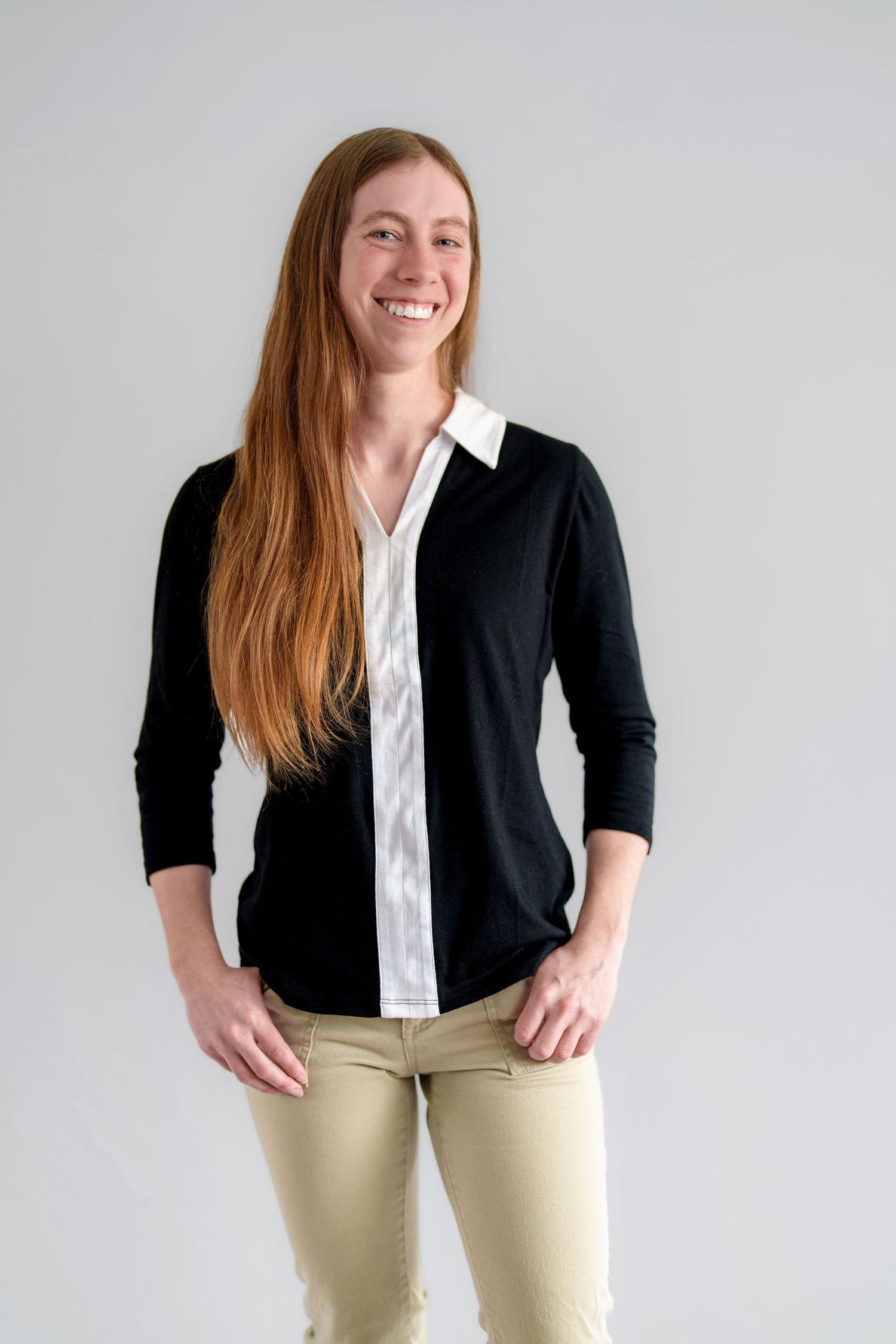 Smiling woman with long red hair wearing a black cardigan over white shirt and khaki pants against gray background.