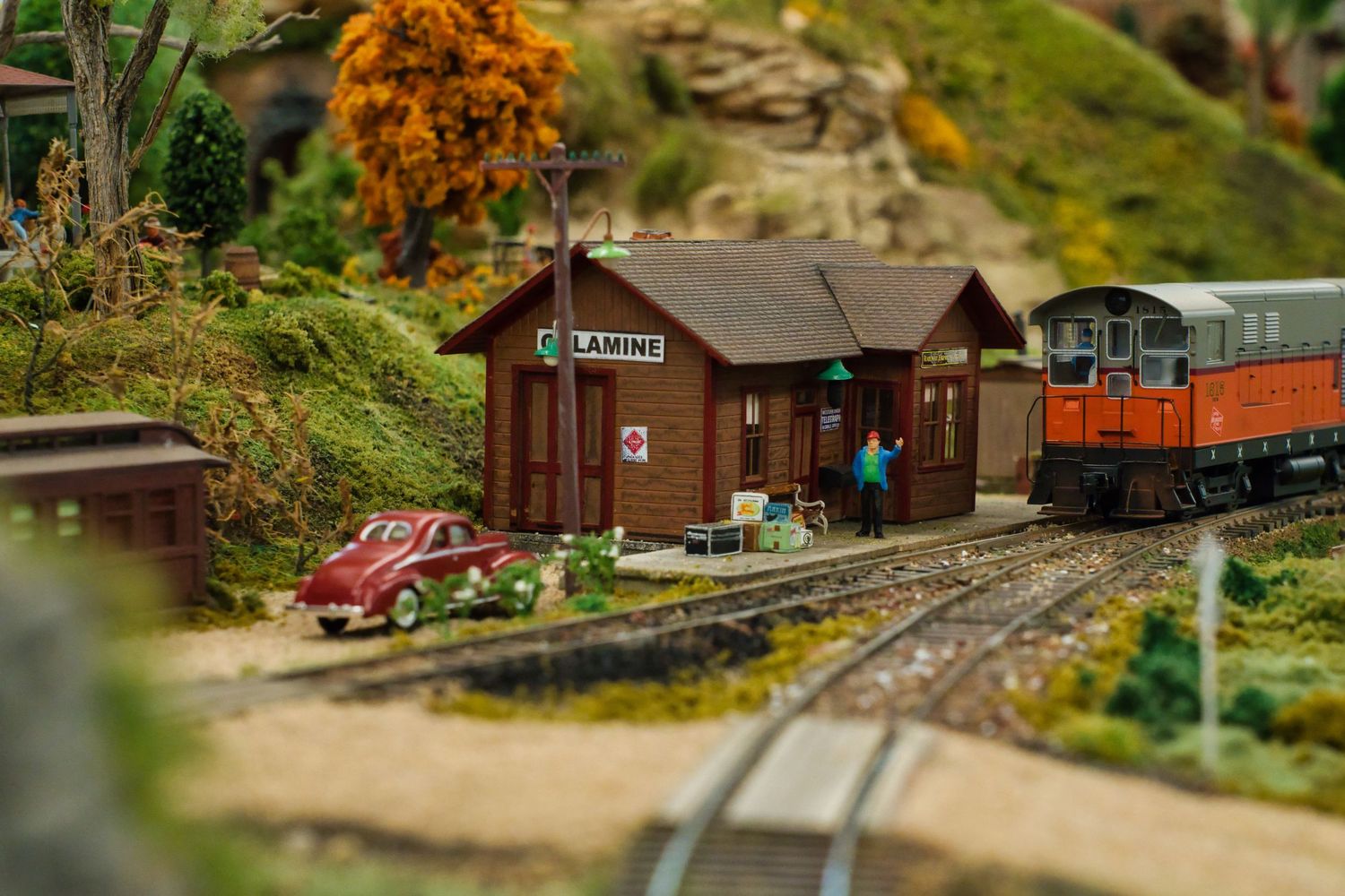 Miniature model train station with brown wooden building, vintage red car, and orange locomotive on detailed railway tracks.