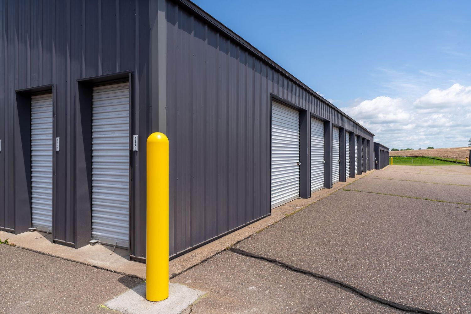 Row of dark gray storage units with yellow safety bollards and gravel parking area under blue sky.