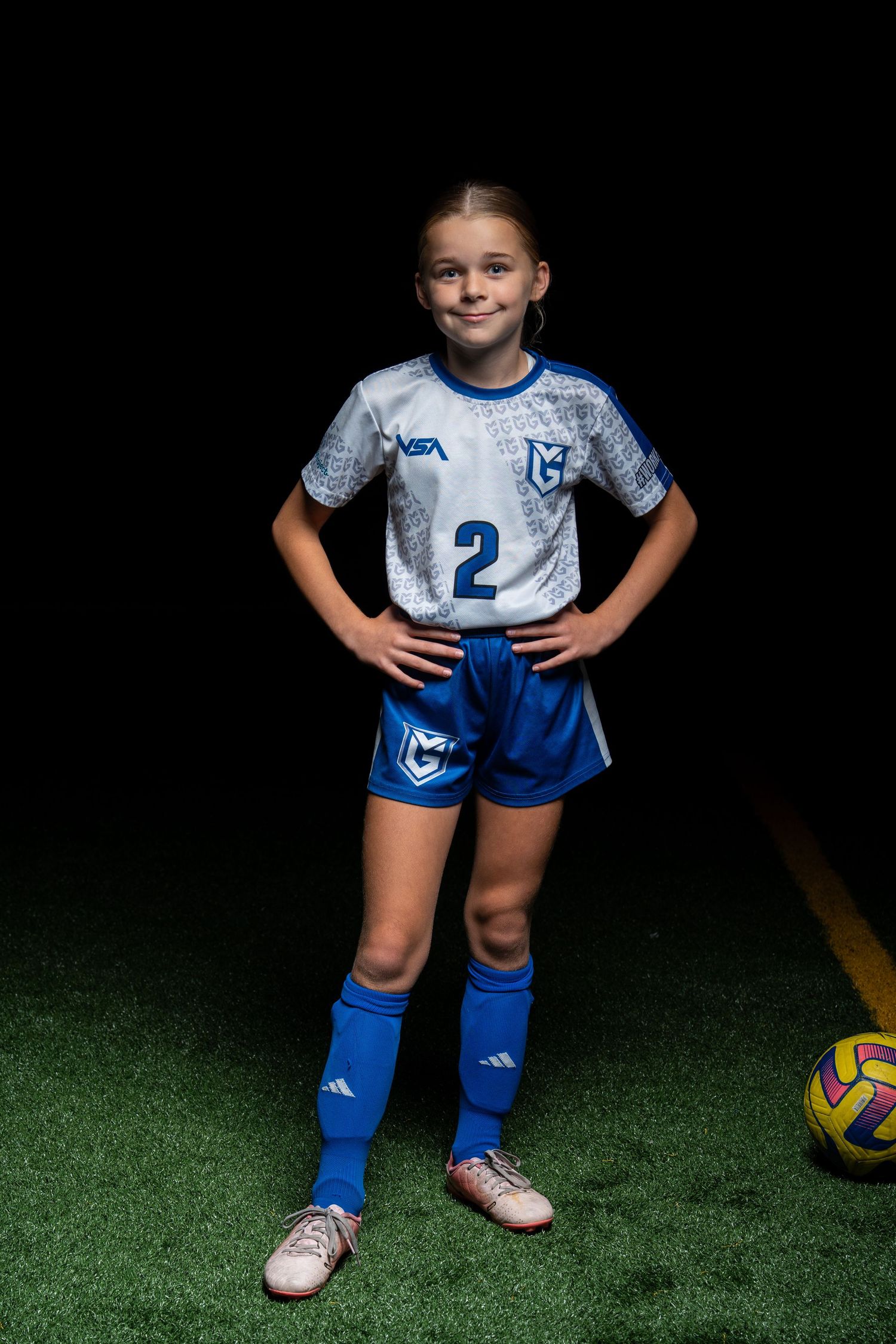 Youth soccer player in blue and white uniform with number 2 poses on artificial turf with soccer ball.