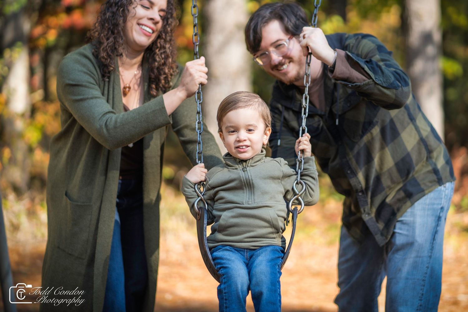 A young child enjoys swinging at a playground on an autumn day with adults helping on either side.