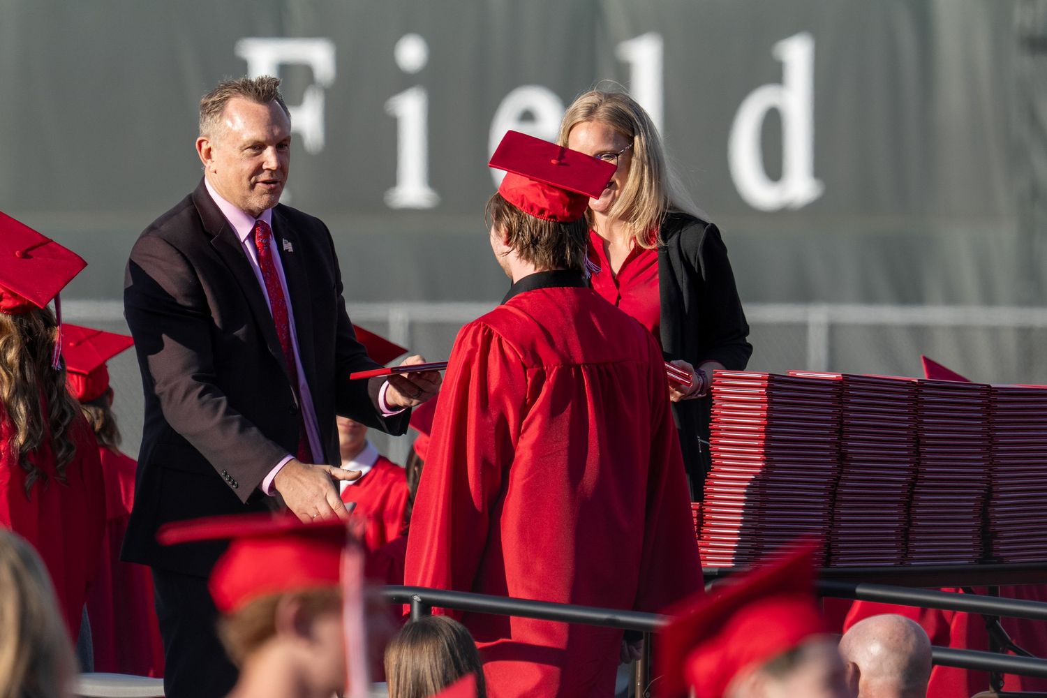Students in red graduation gowns receive diplomas on stage at an outdoor commencement ceremony.
