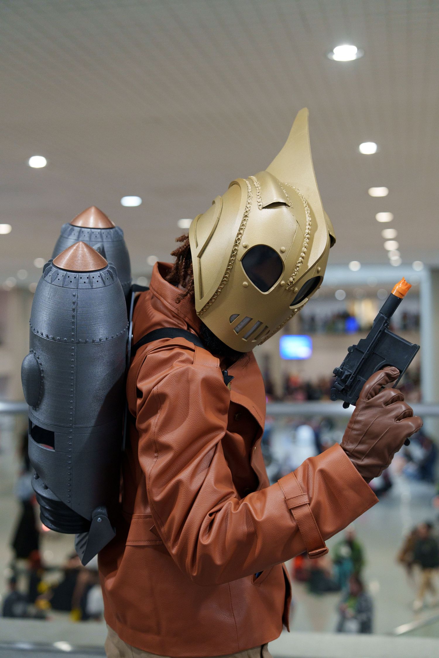 Person cosplaying as The Rocketeer with golden helmet, rocket pack, and toy gun at a convention.