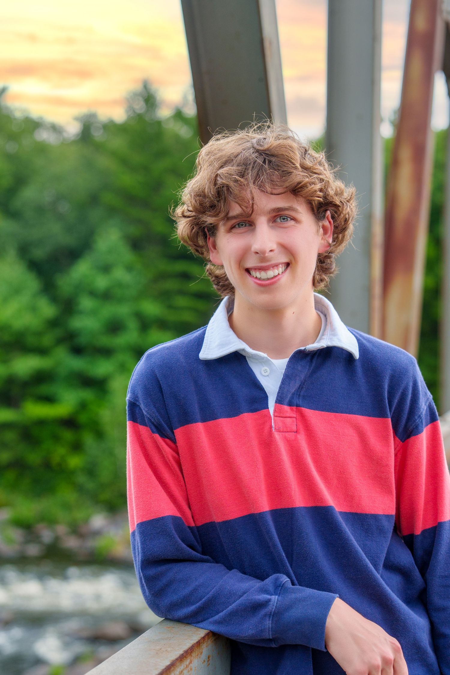 Smiling teenage boy with curly hair wearing a blue and red striped polo shirt outdoors at sunset.