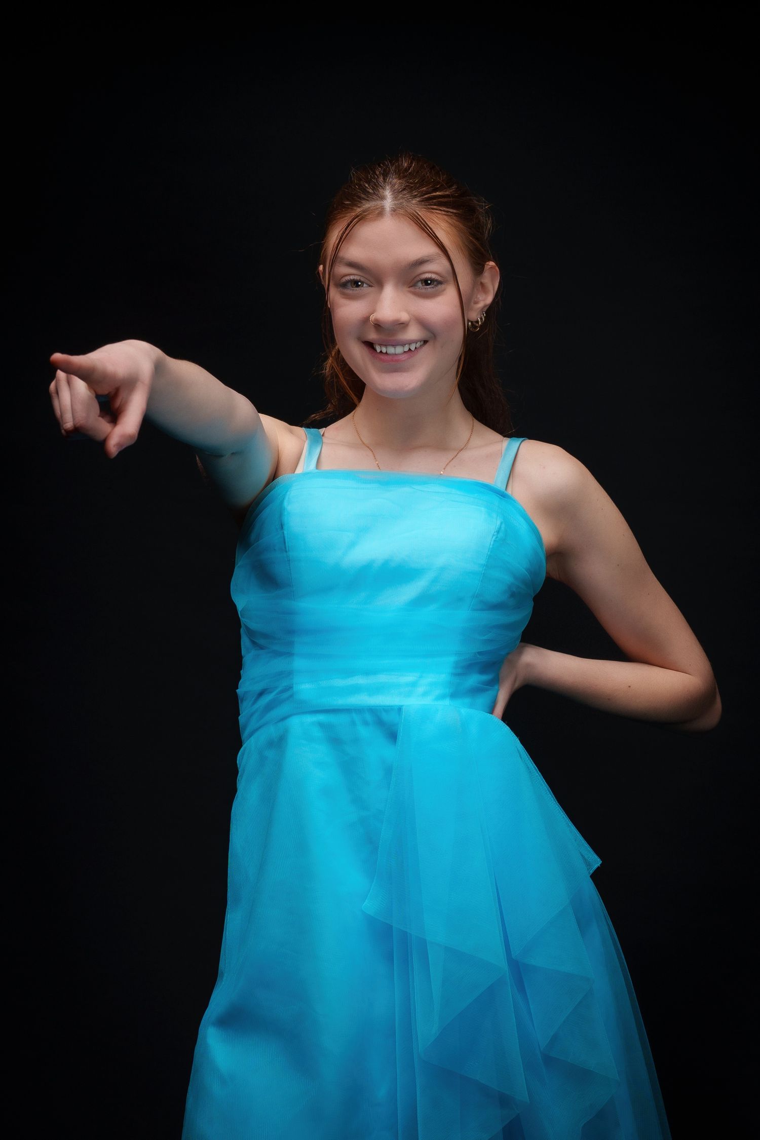 Smiling teenage girl in a blue tulle dress pointing forward against a dark background.