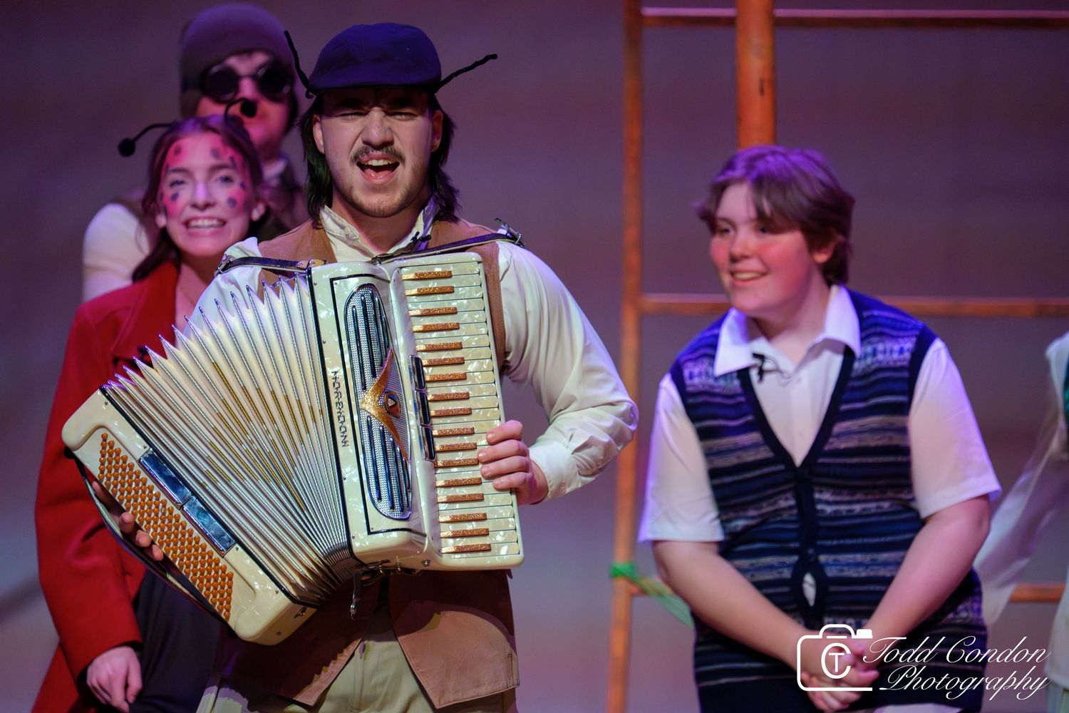 Musicians perform on stage with an accordion player in the center wearing a baseball cap during a theatrical production.