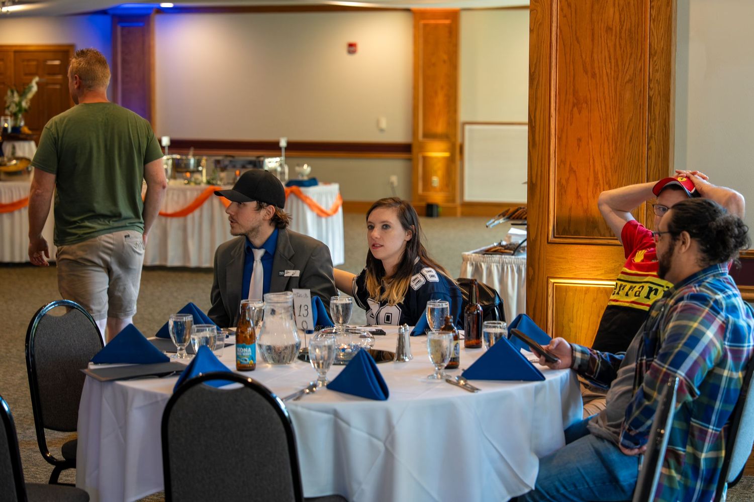 People gathered around a formal table with blue napkins and place settings in a banquet room with wood accents.