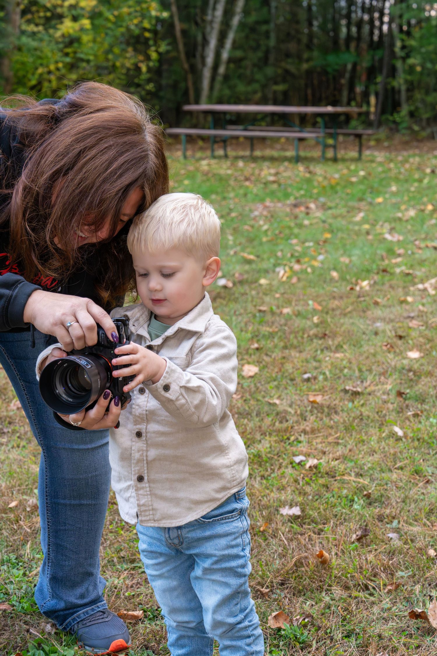 A woman bends down to show a young blonde toddler boy how to use a DSLR camera outdoors in a park.