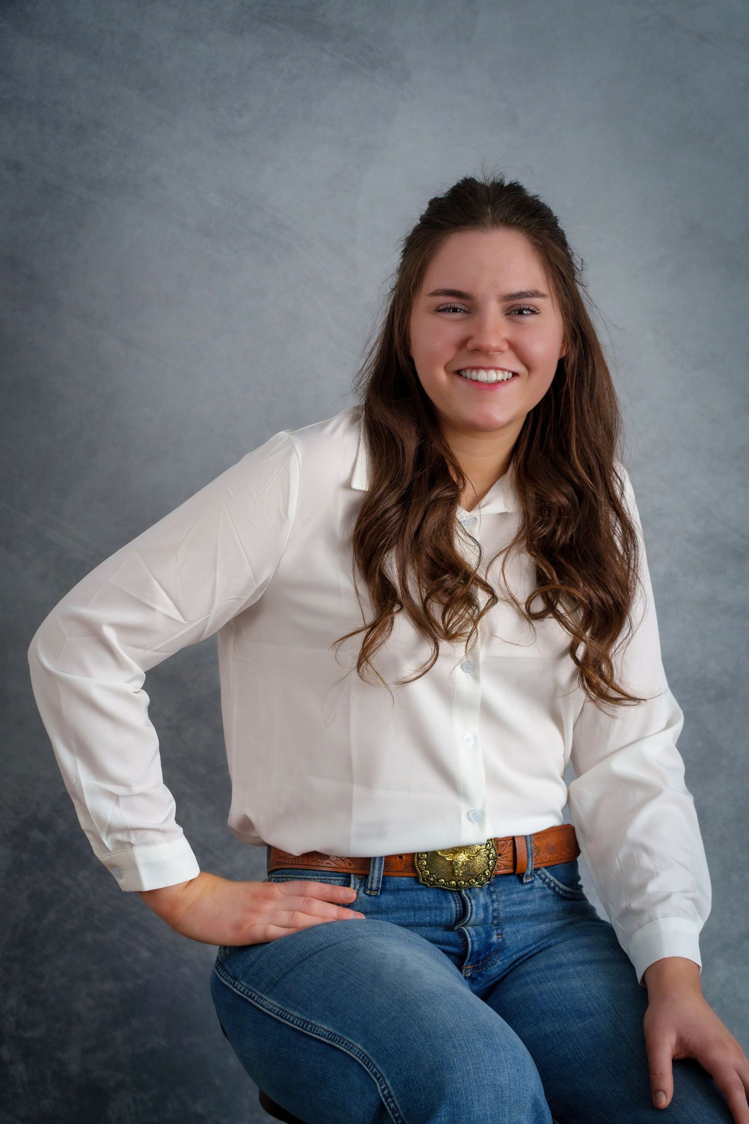 Smiling young woman in white button-up shirt and jeans with decorative belt, posing for a portrait photo.