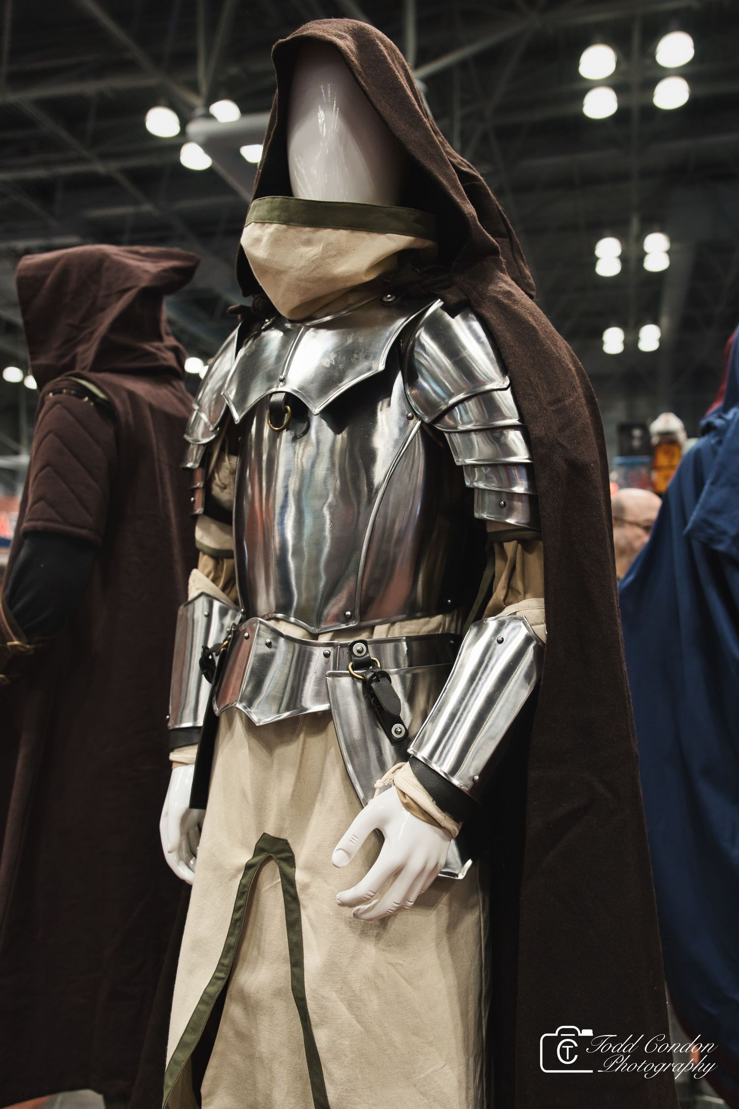 A mannequin dressed in medieval knight armor with a brown hooded cloak on display at a convention.