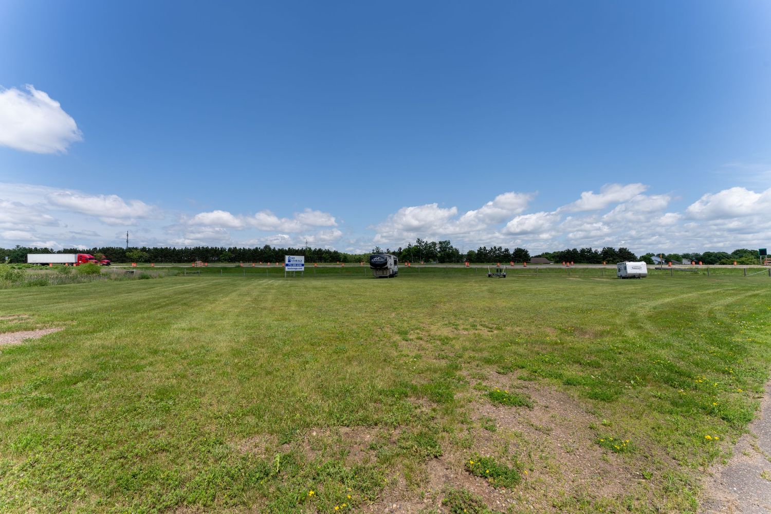Panoramic view of a vast green field with dirt paths and scattered trees under a partly cloudy blue sky.