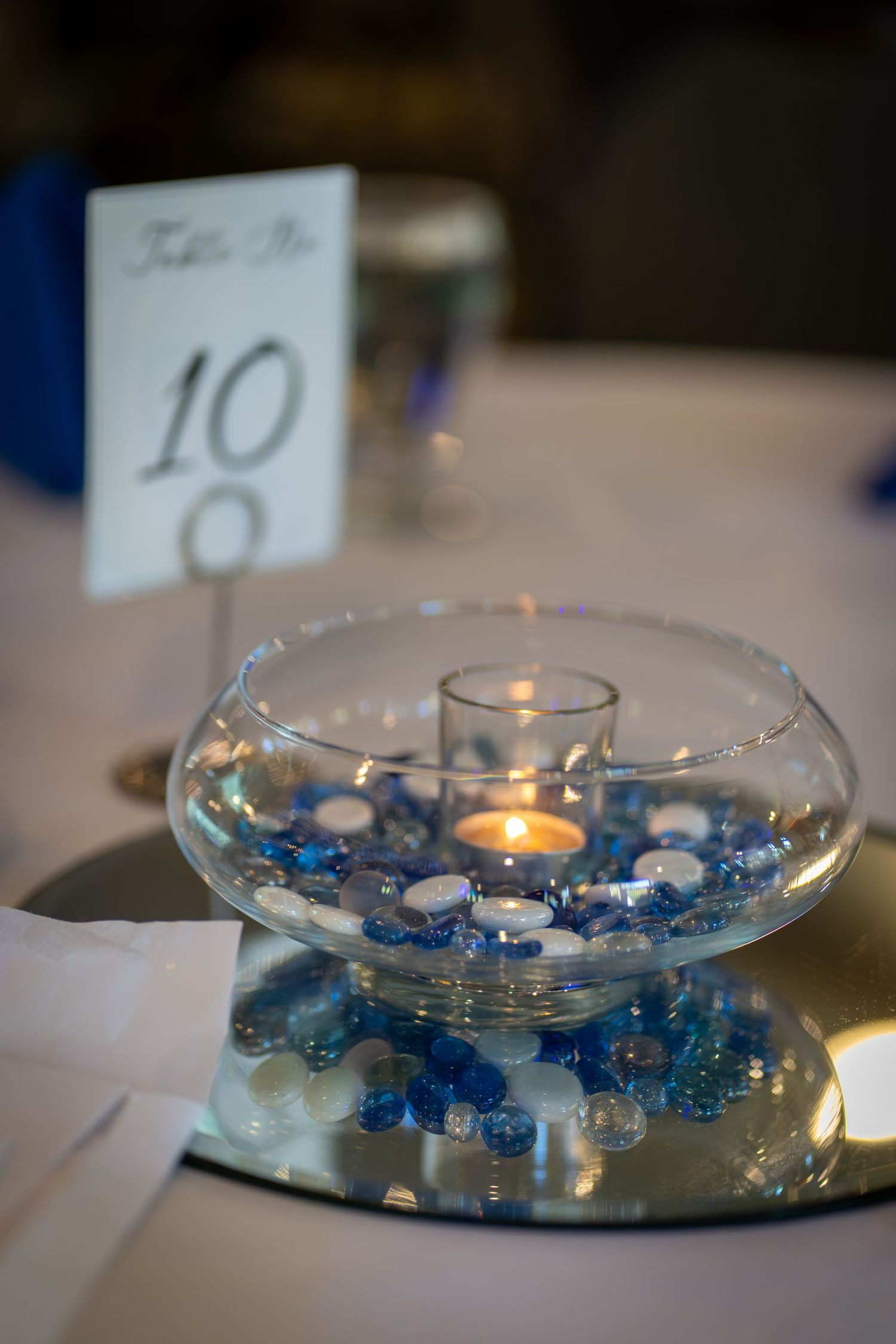 Glass candle centerpiece with blue and clear decorative stones glowing softly on a mirrored surface.