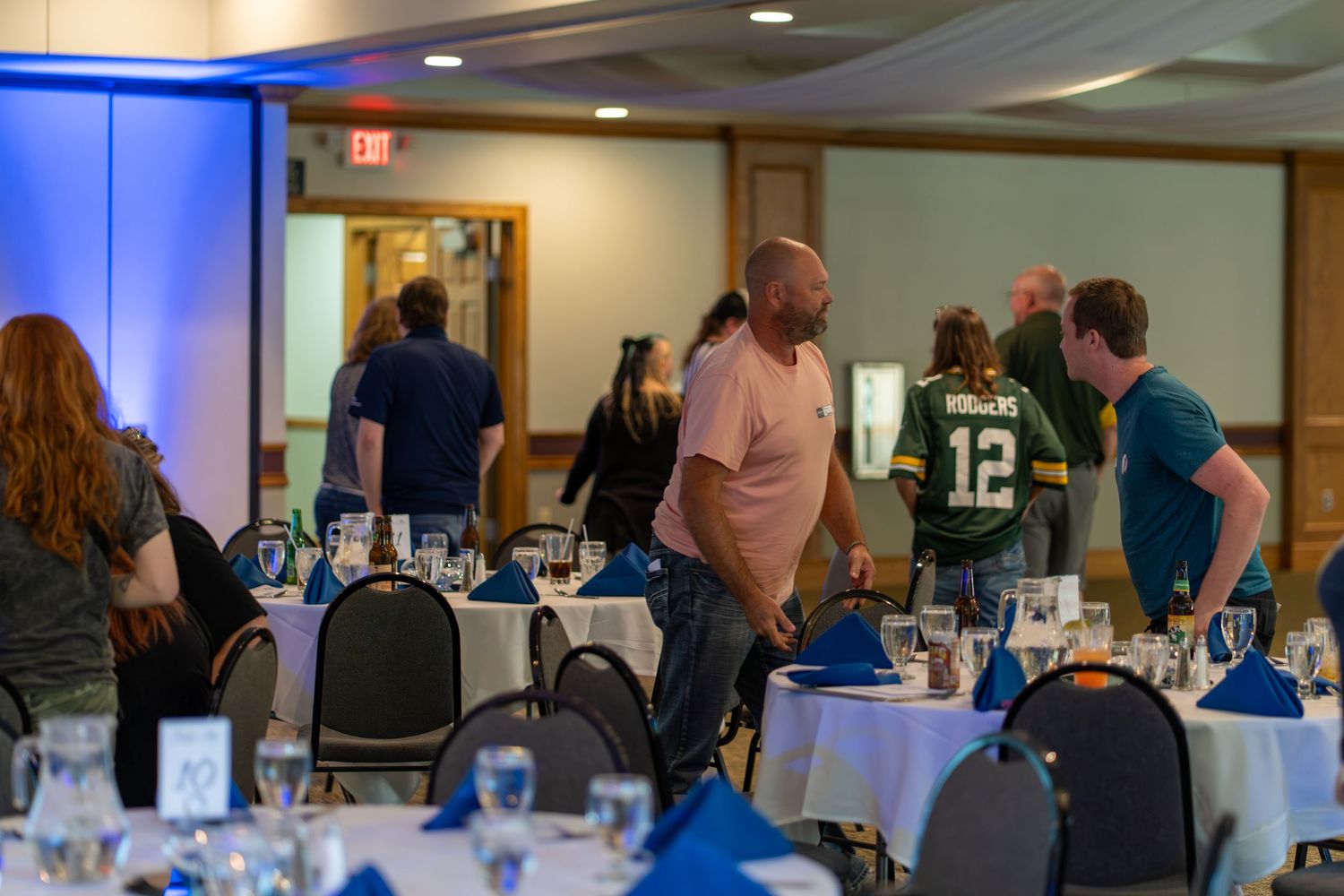 People gather at a social event in a room with blue lighting and tables set up for a meeting or conference.