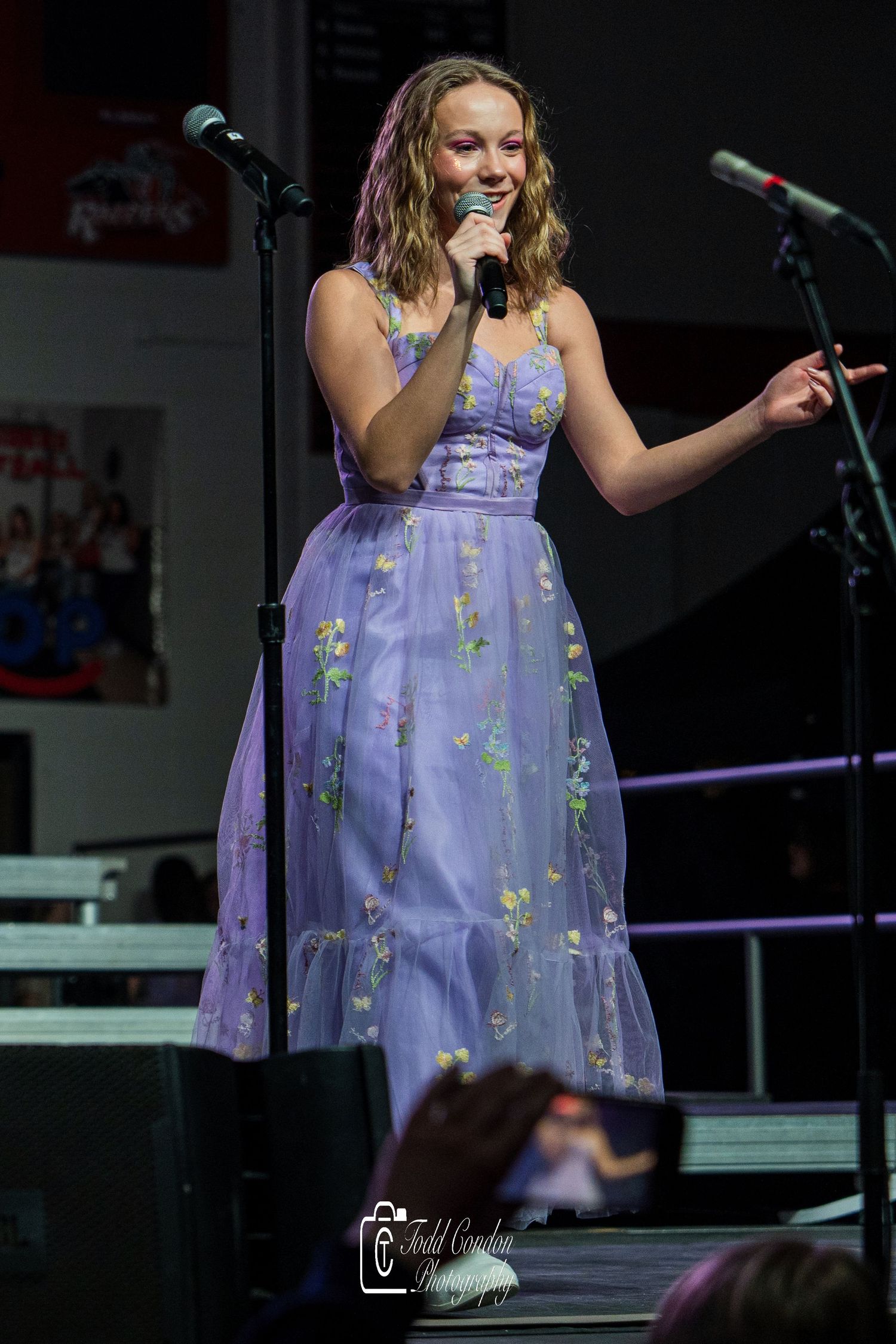 A performer in a purple floral dress sings on stage during a live performance.