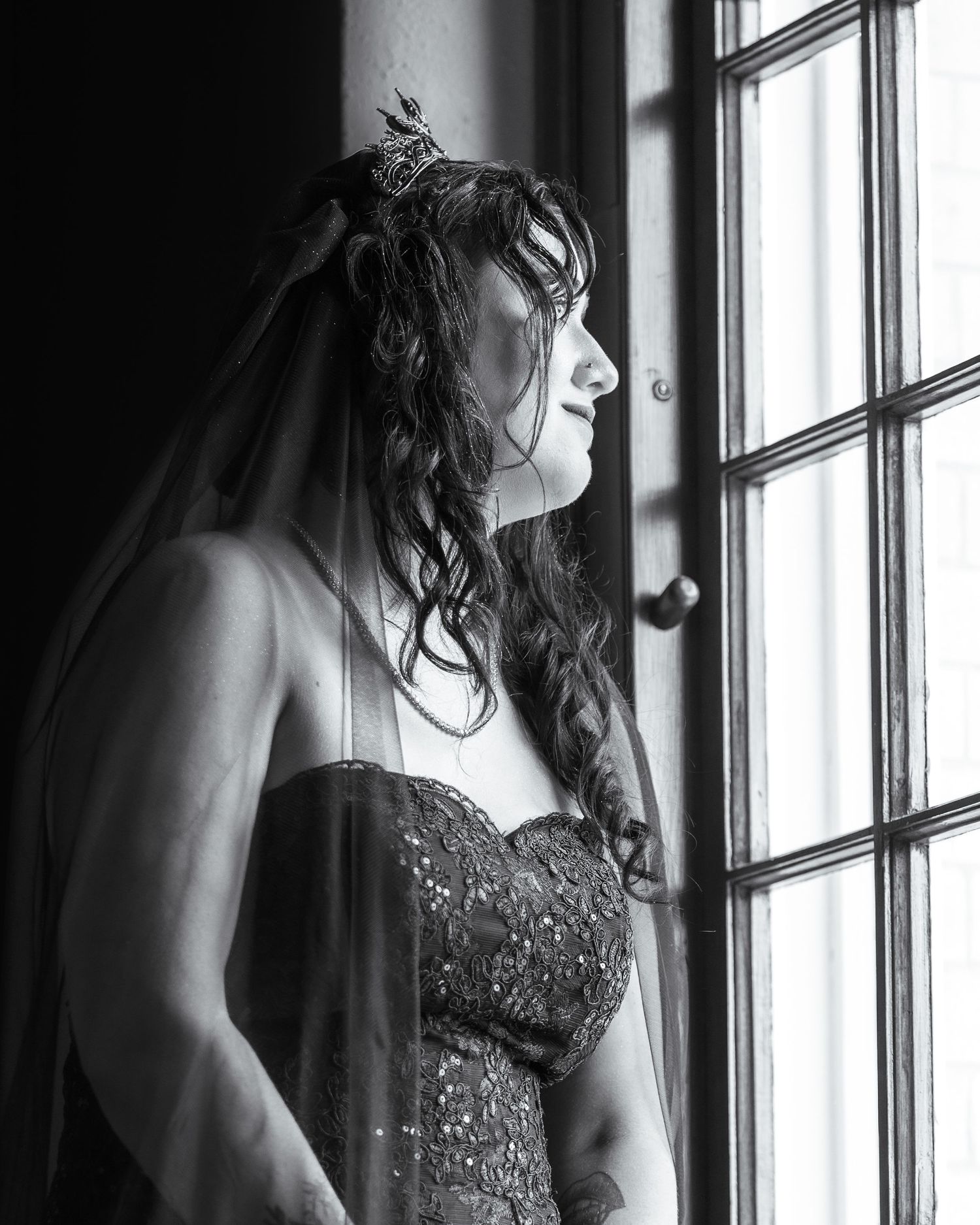 A black and white portrait of a woman in an elegant dress gazing through a window in dramatic lighting.