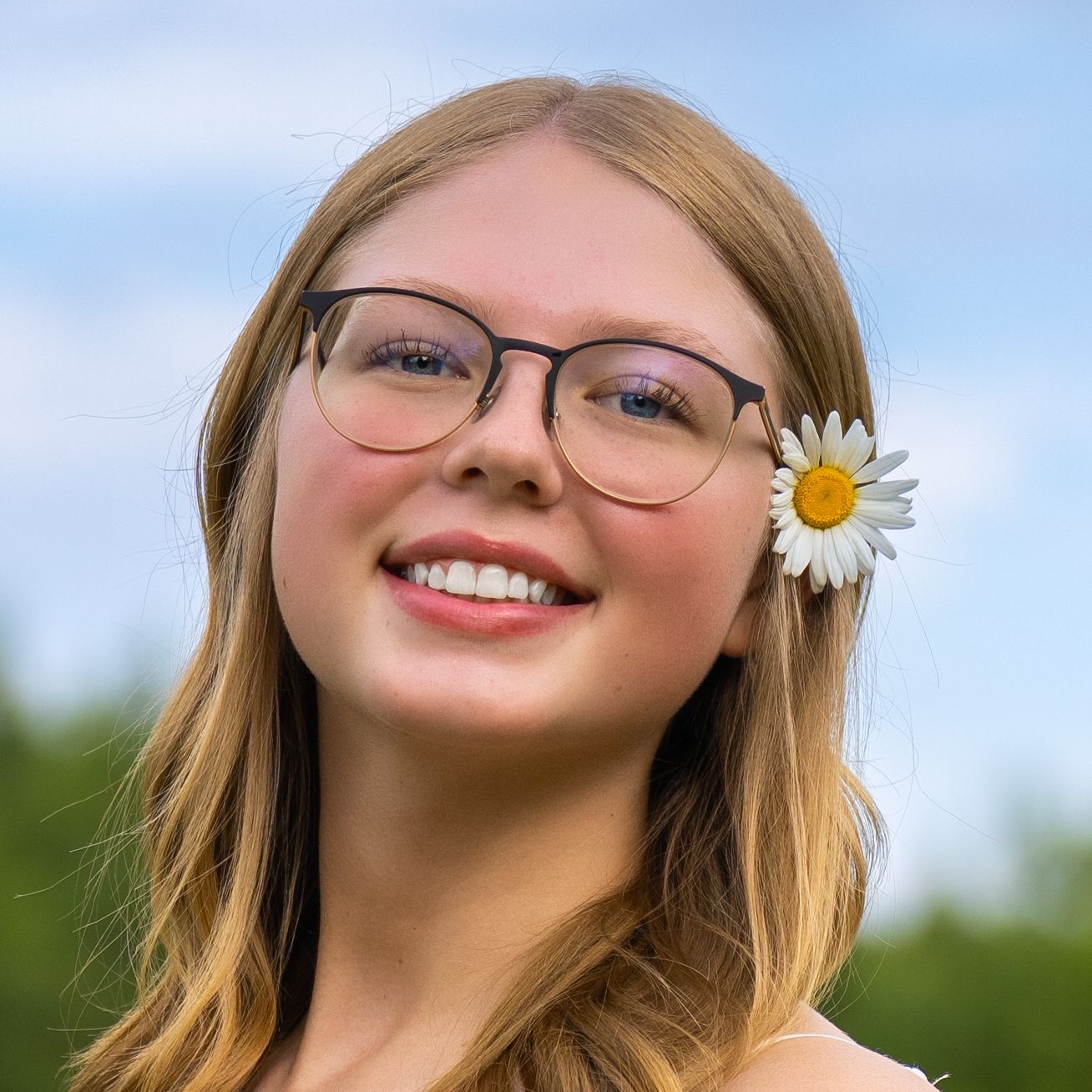 Person with glasses and blonde hair smiling outdoors with a white daisy flower tucked behind their ear in sunlight.