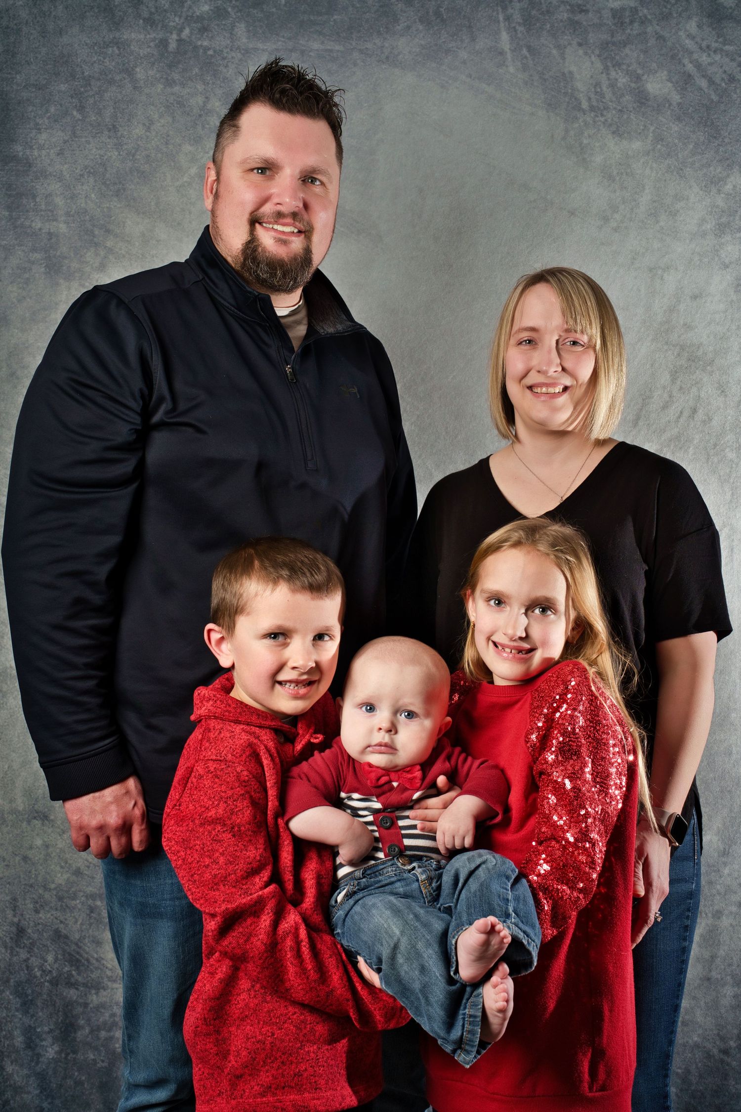 A family portrait with parents and three children wearing red holiday attire against a gray background.