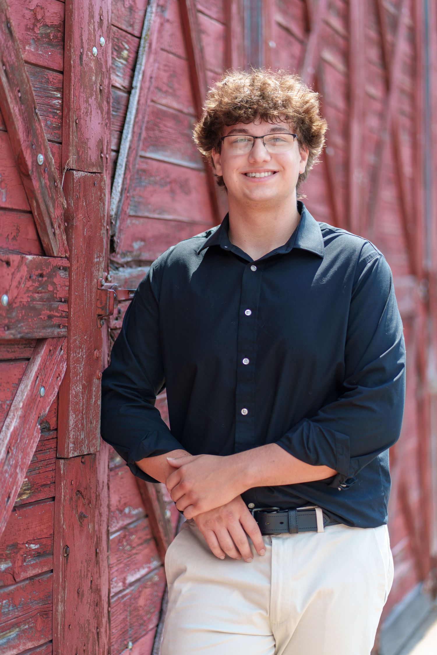 Smiling teenage boy with curly hair and glasses in navy shirt leaning against rustic red barn wall.