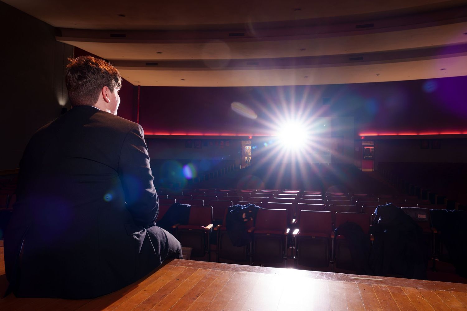 Person sitting alone in an empty theater auditorium with bright stage spotlight illuminating red velvet seats.