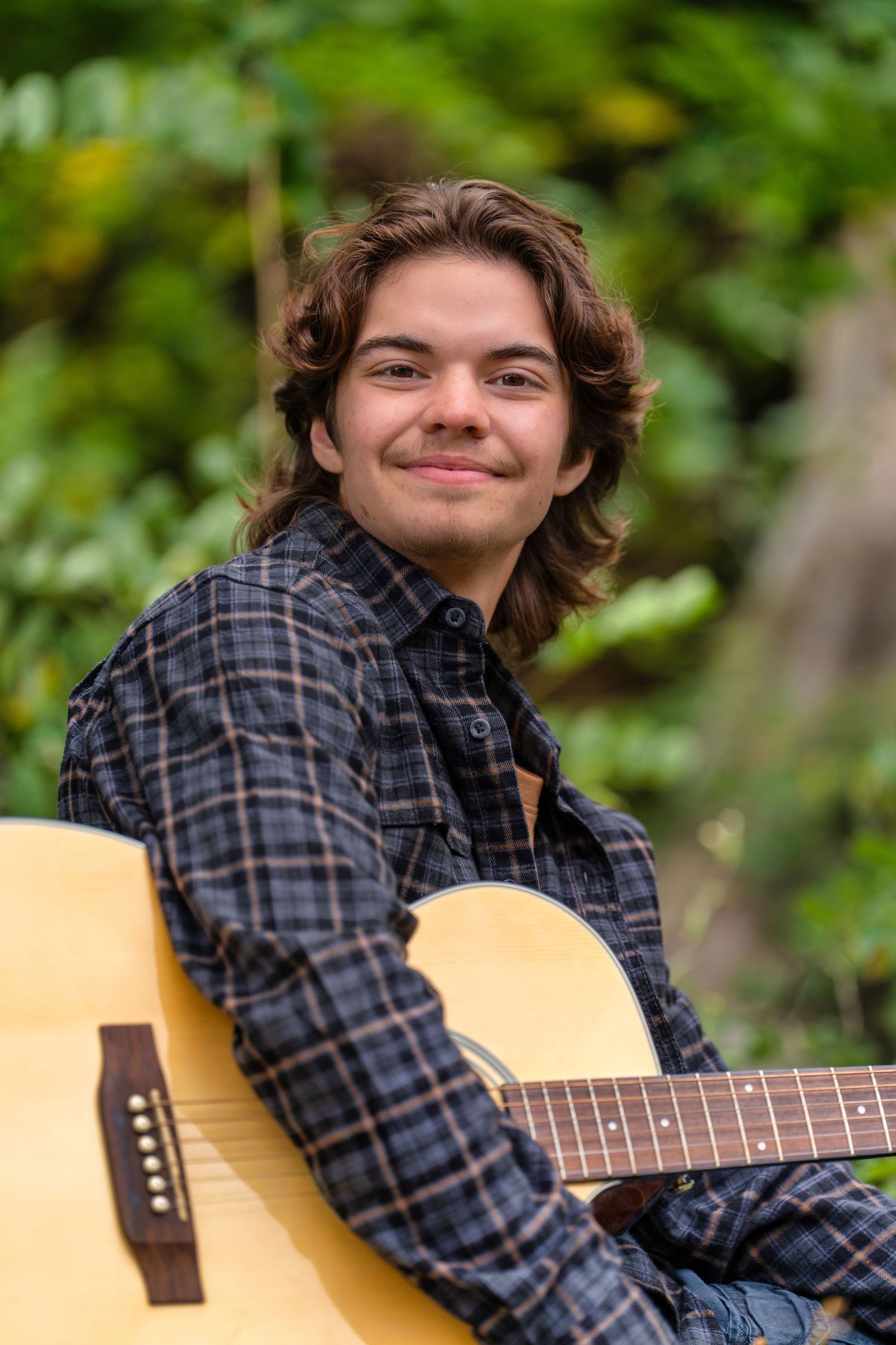 A person wearing a plaid flannel shirt plays an acoustic guitar outdoors with greenery in the background.