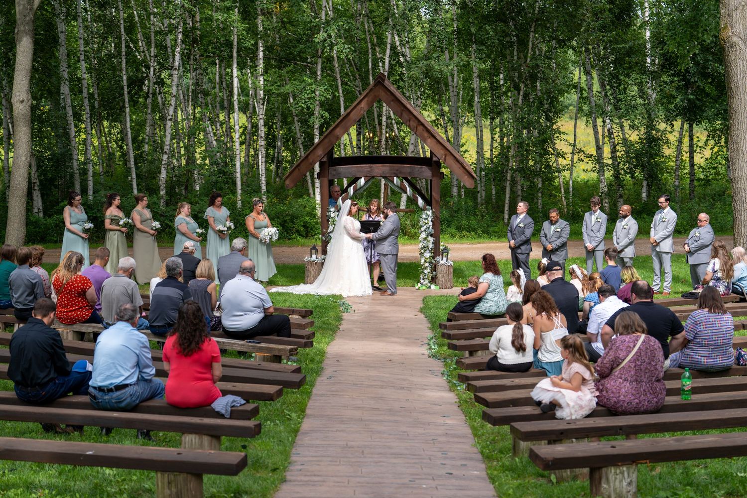 People sit on wooden benches in a tranquil outdoor amphitheater surrounded by lush green trees.