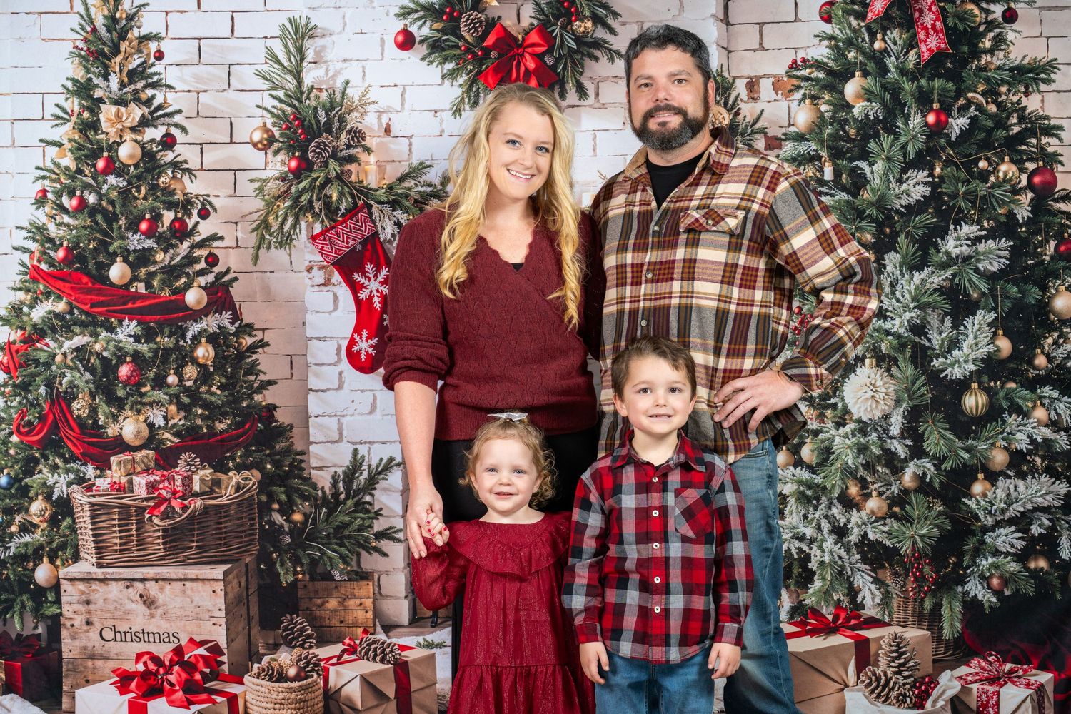 Family wearing coordinating red and plaid outfits poses for Christmas photos in front of white brick wall and trees.