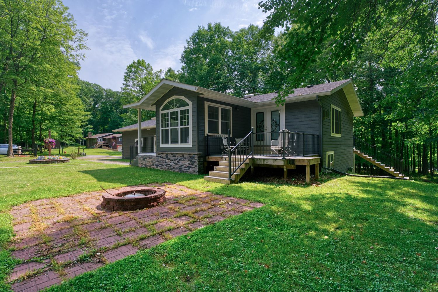 Gray cabin with brick accents, wooden deck, fire pit patio, and lush green lawn surrounded by tall trees.