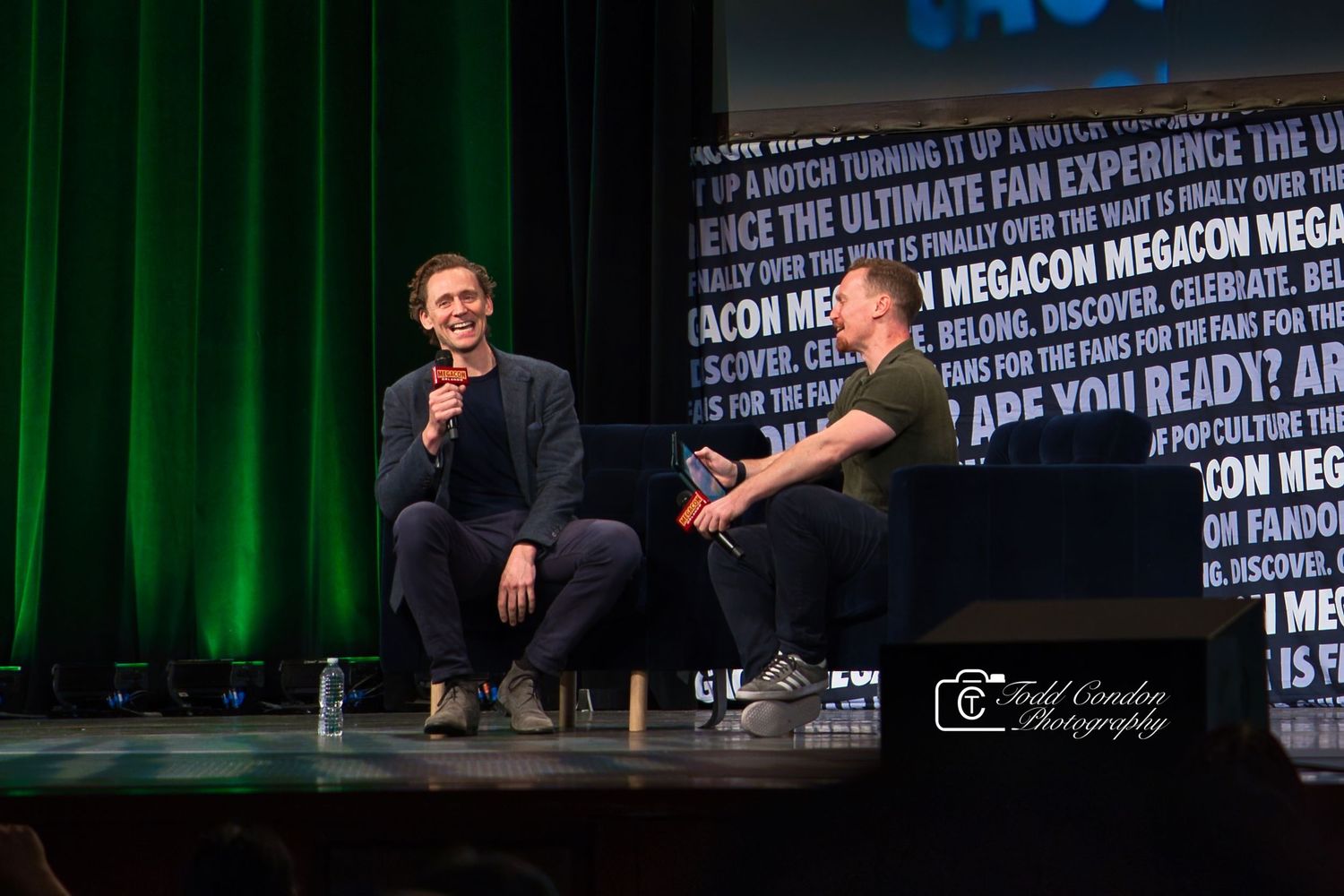 Speakers engage in discussion on stage at a technology conference with green lighting and text projections on wall.