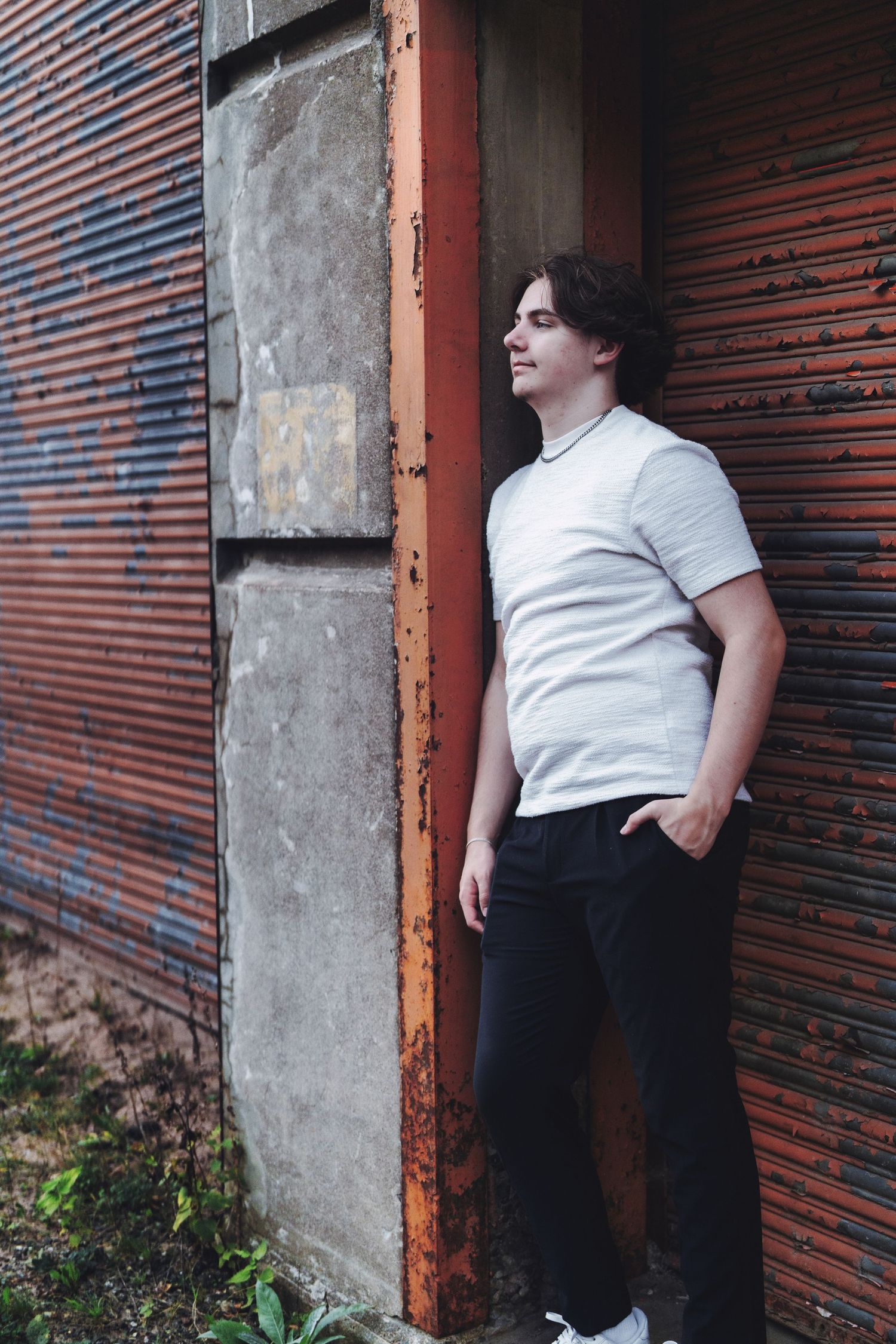 A person in a white shirt and dark pants leans against a concrete wall near a metal shutter door.