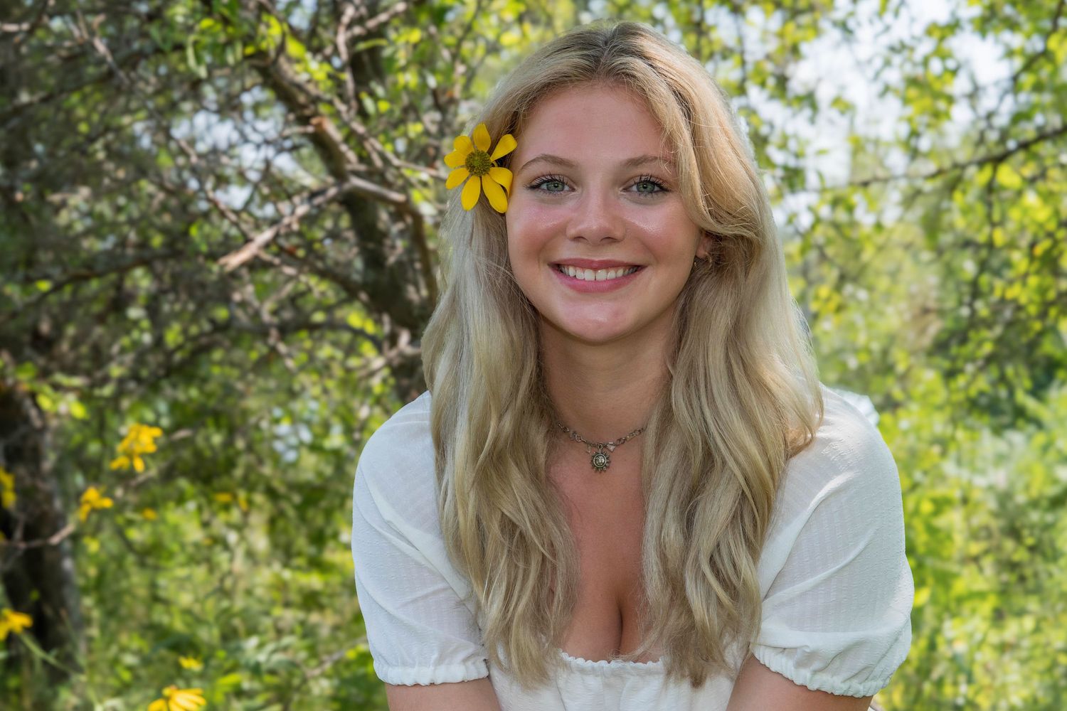 A person with blonde hair smiles warmly while sitting outdoors among green foliage and yellow flowers in natural lighting.