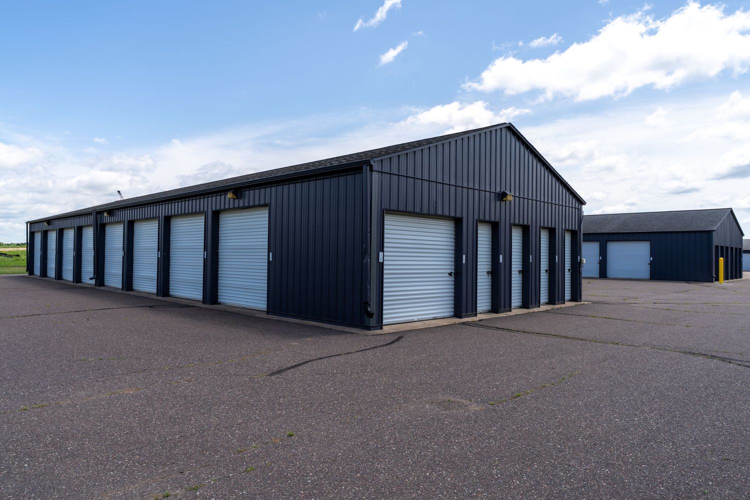 Exterior view of modern black metal self-storage units with roll-up doors arranged in rows under a blue sky.