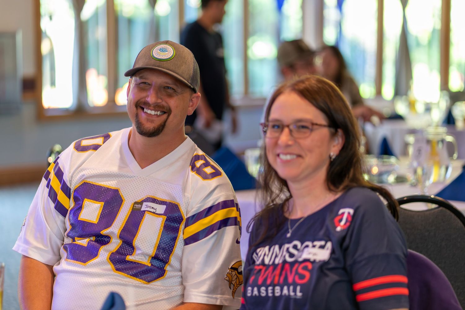 Two sports fans wearing team jerseys smile together at an indoor event.