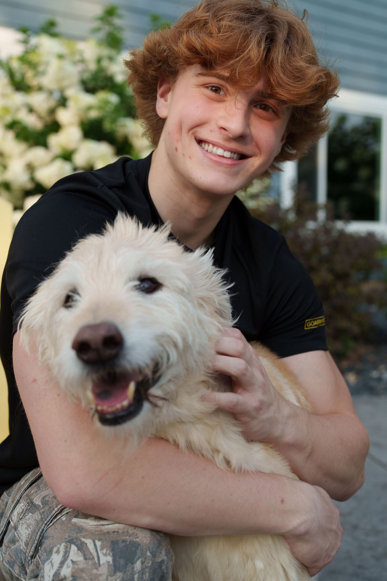 Smiling young man with curly red hair hugging a fluffy white dog outdoors near a house.