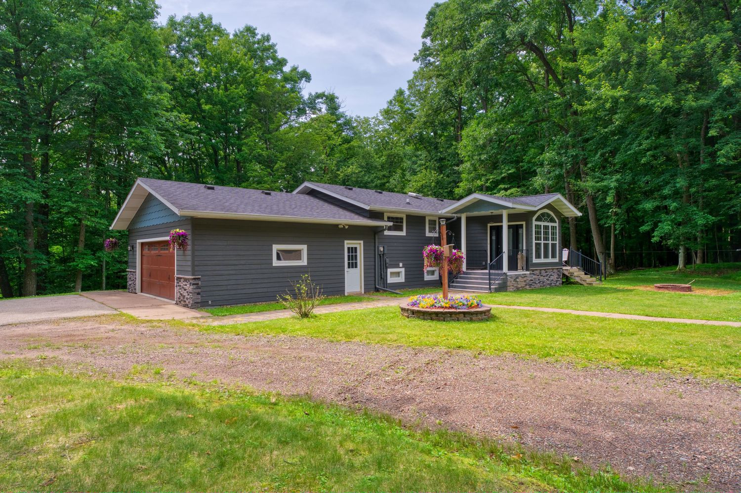 Gray ranch-style home with red garage door surrounded by lush green trees and manicured lawn in a wooded setting.