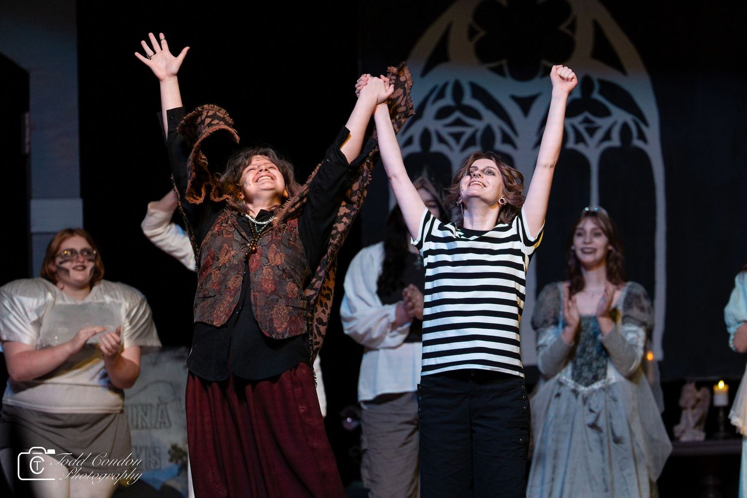 Performers on stage raise their arms in celebration during a theatrical performance with dark lighting and dramatic staging.