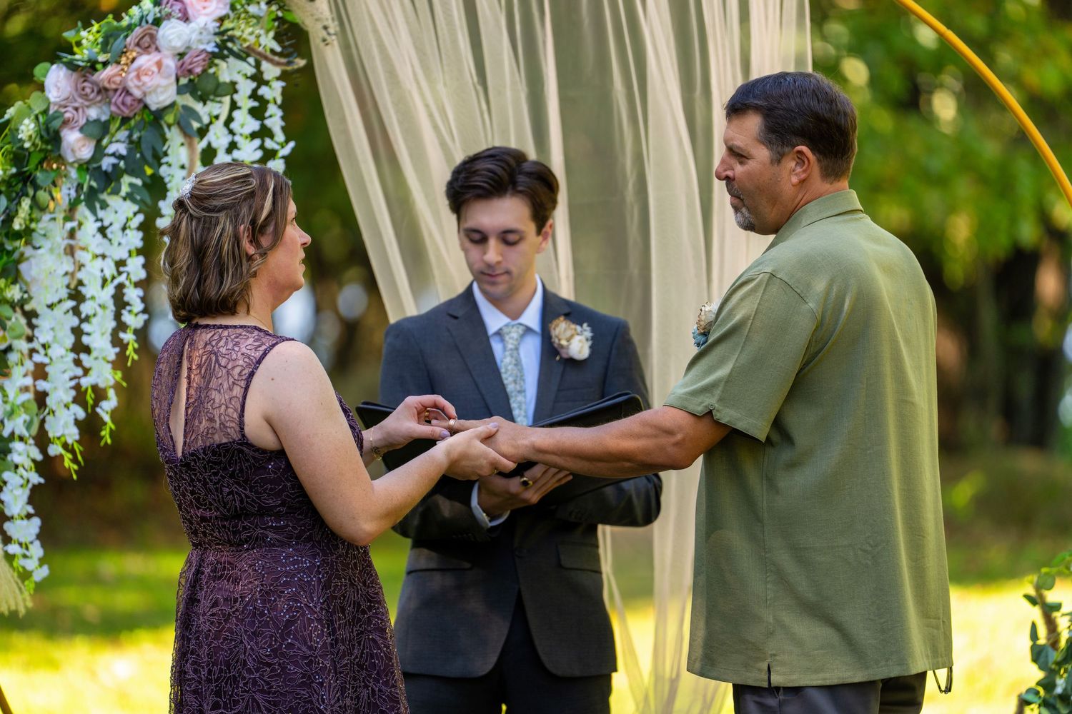 A wedding ceremony takes place outdoors under a floral arch with wisteria and white fabric draping.
