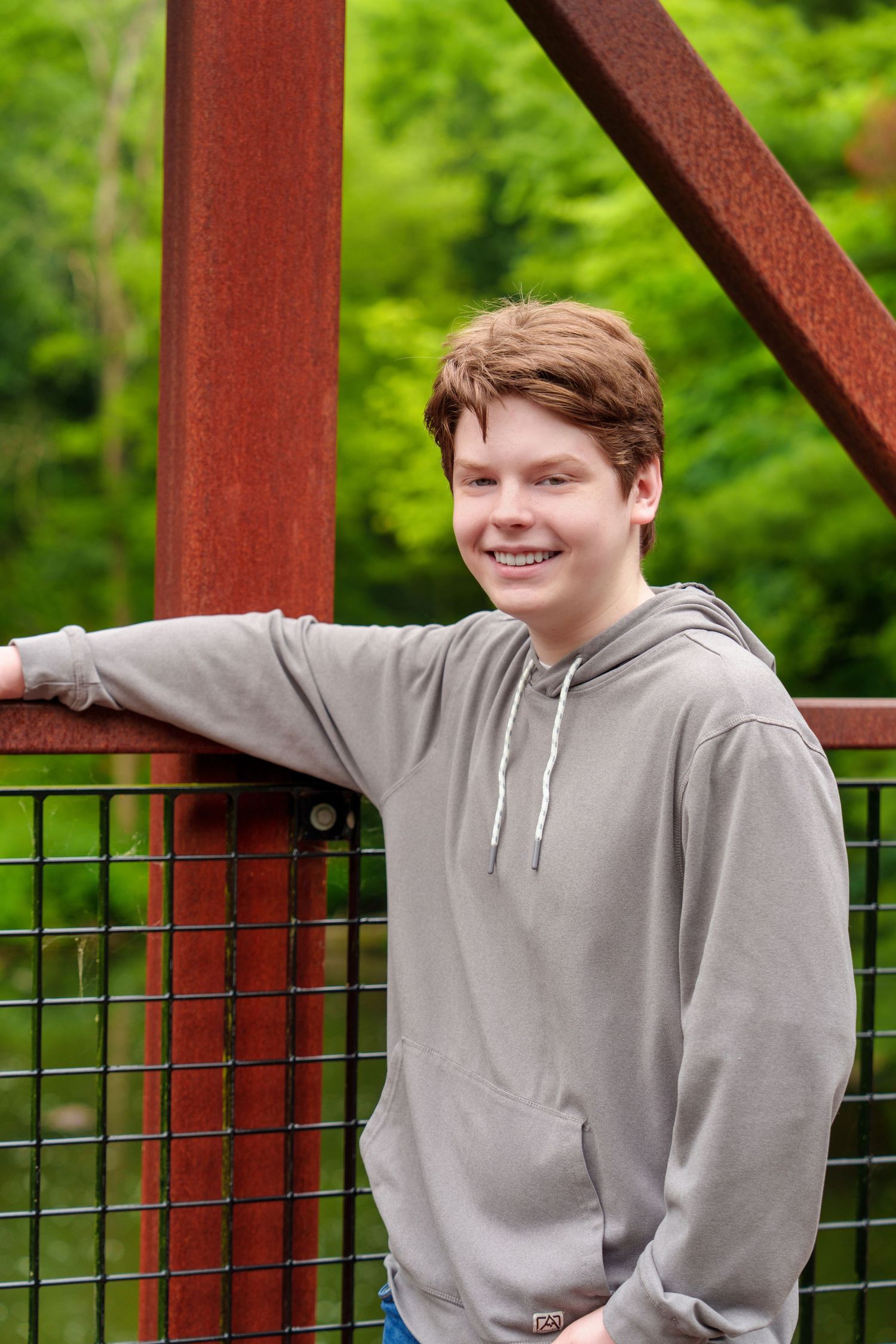 Smiling teenage boy in gray hoodie leaning on rusty red bridge railing with lush green trees in background.