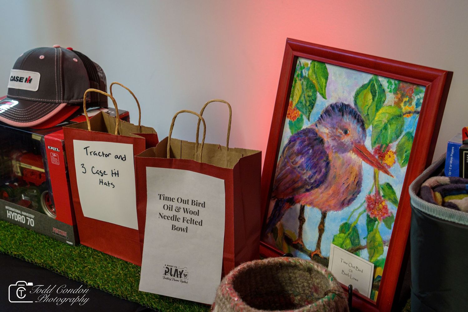 Colorful needle felted bird artwork in red frame displayed at art show beside brown paper bags and a woven basket.