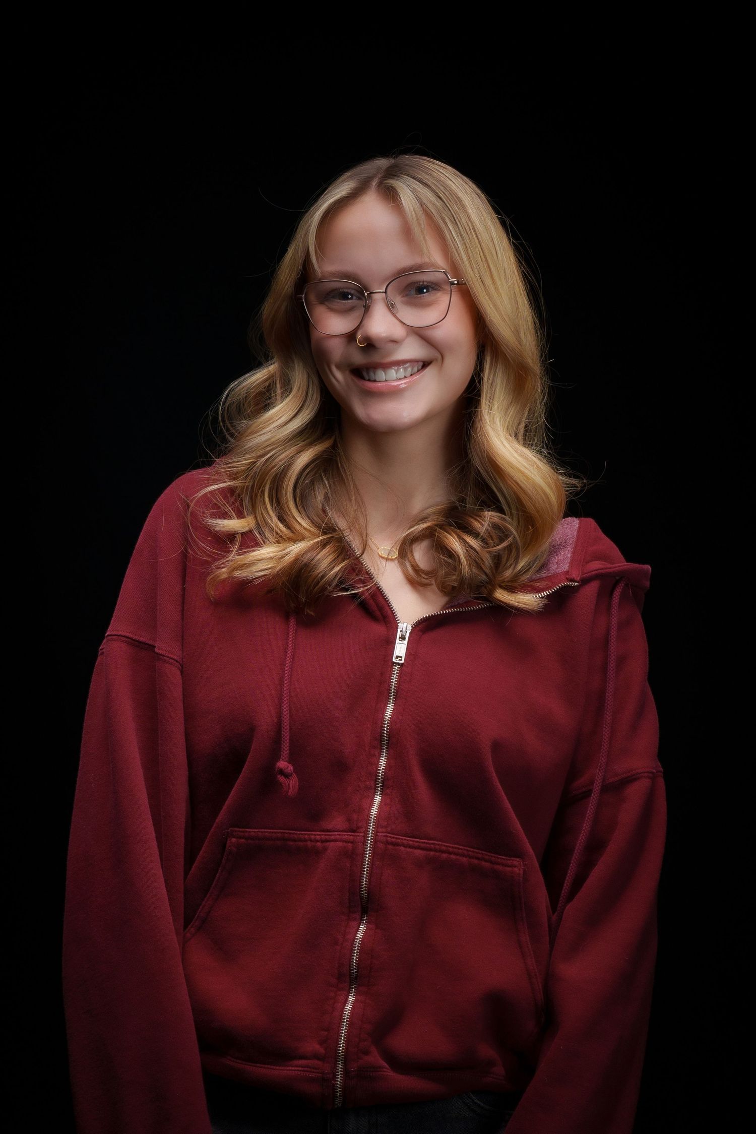 Smiling teenage girl with blonde hair, glasses, and braces wearing a red zip-up hoodie against a black background.