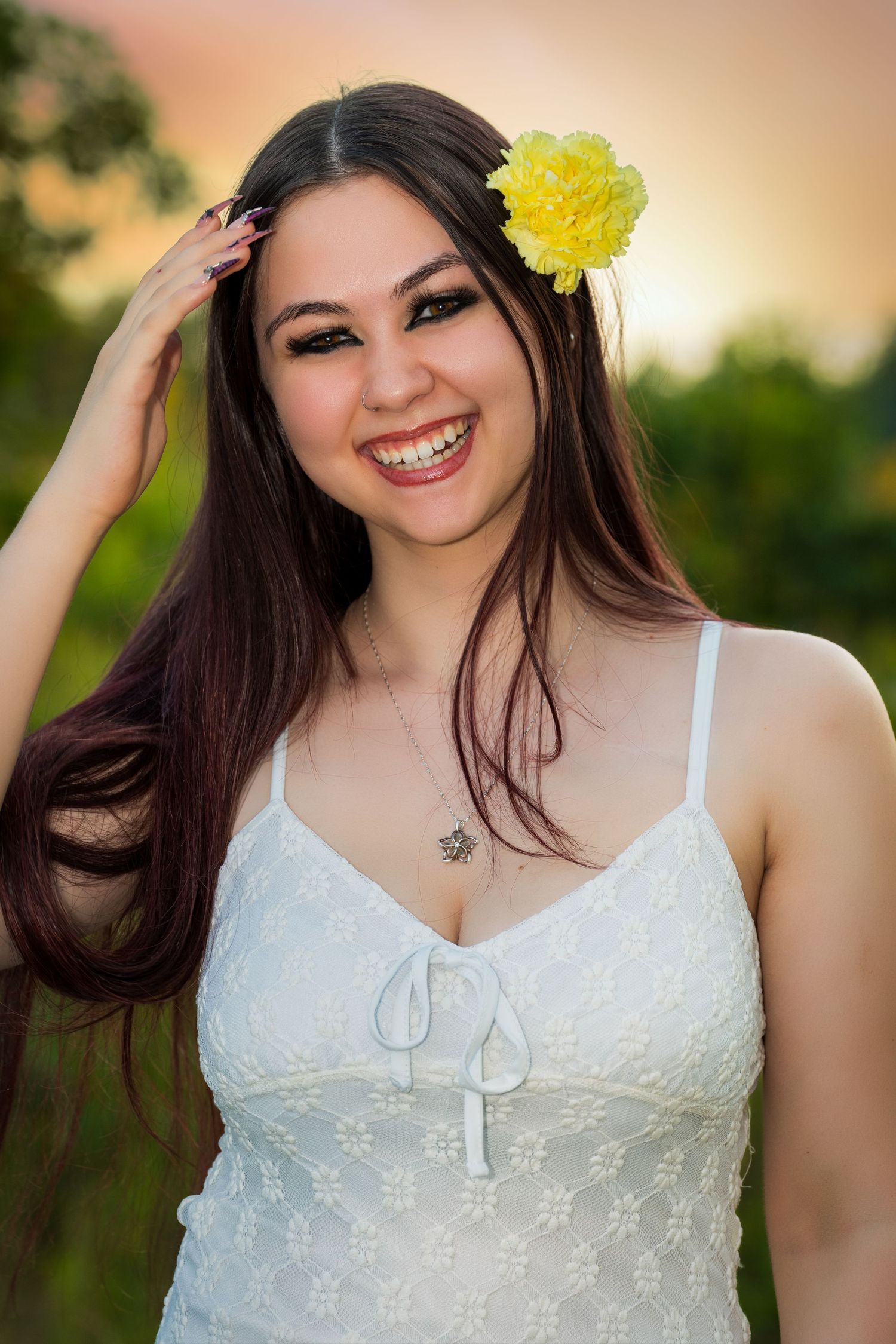 Smiling young woman with long dark hair, yellow flower accessory, wearing white sundress outdoors at sunset.