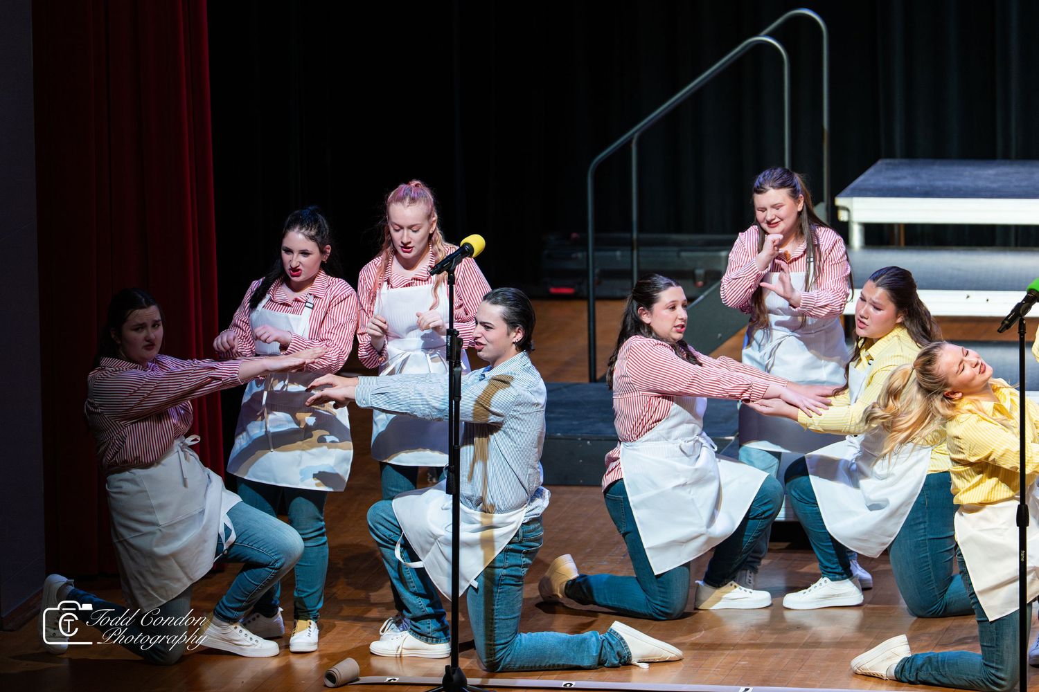Performers in white aprons dance and interact energetically during a theatrical stage production.