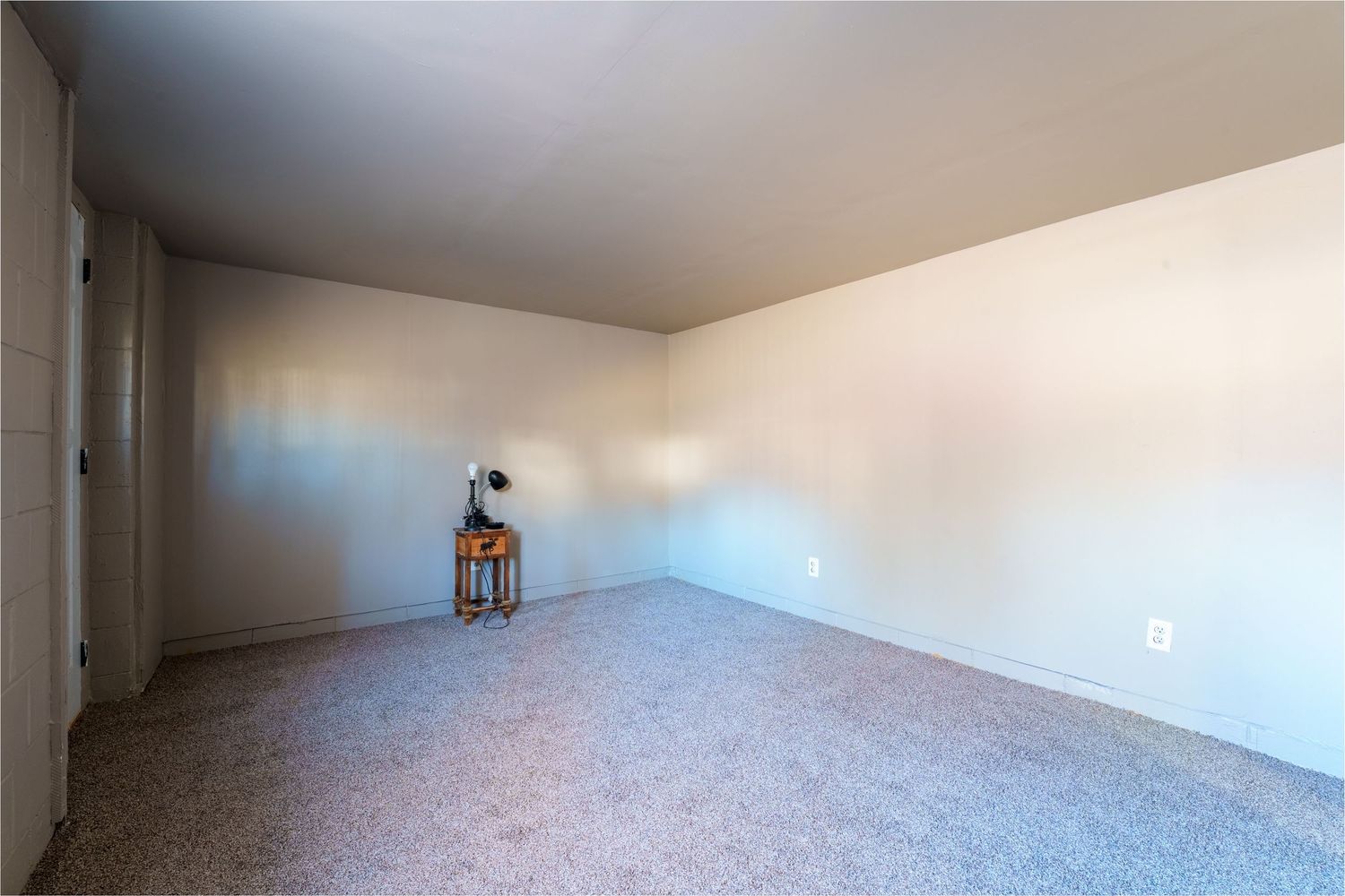 Wide angle view of an empty carpeted basement room with white walls and neutral lighting.