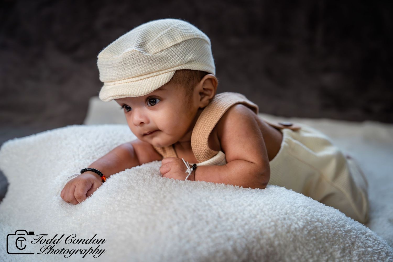 A newborn baby wearing a white hat rests peacefully on soft white textured bedding in a black and white photo.