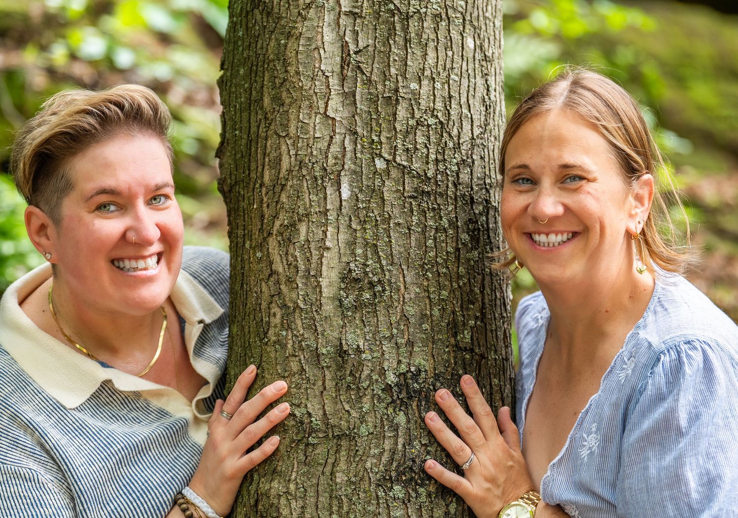 Two people playfully posing against a large tree trunk in a forest setting.