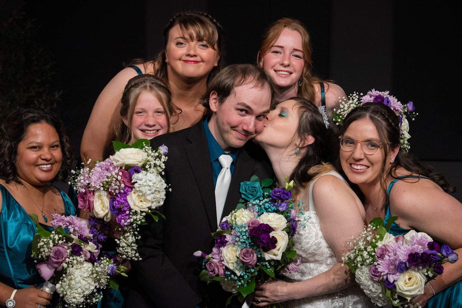 A joyful wedding party poses together with purple and white floral bouquets against a dark background.