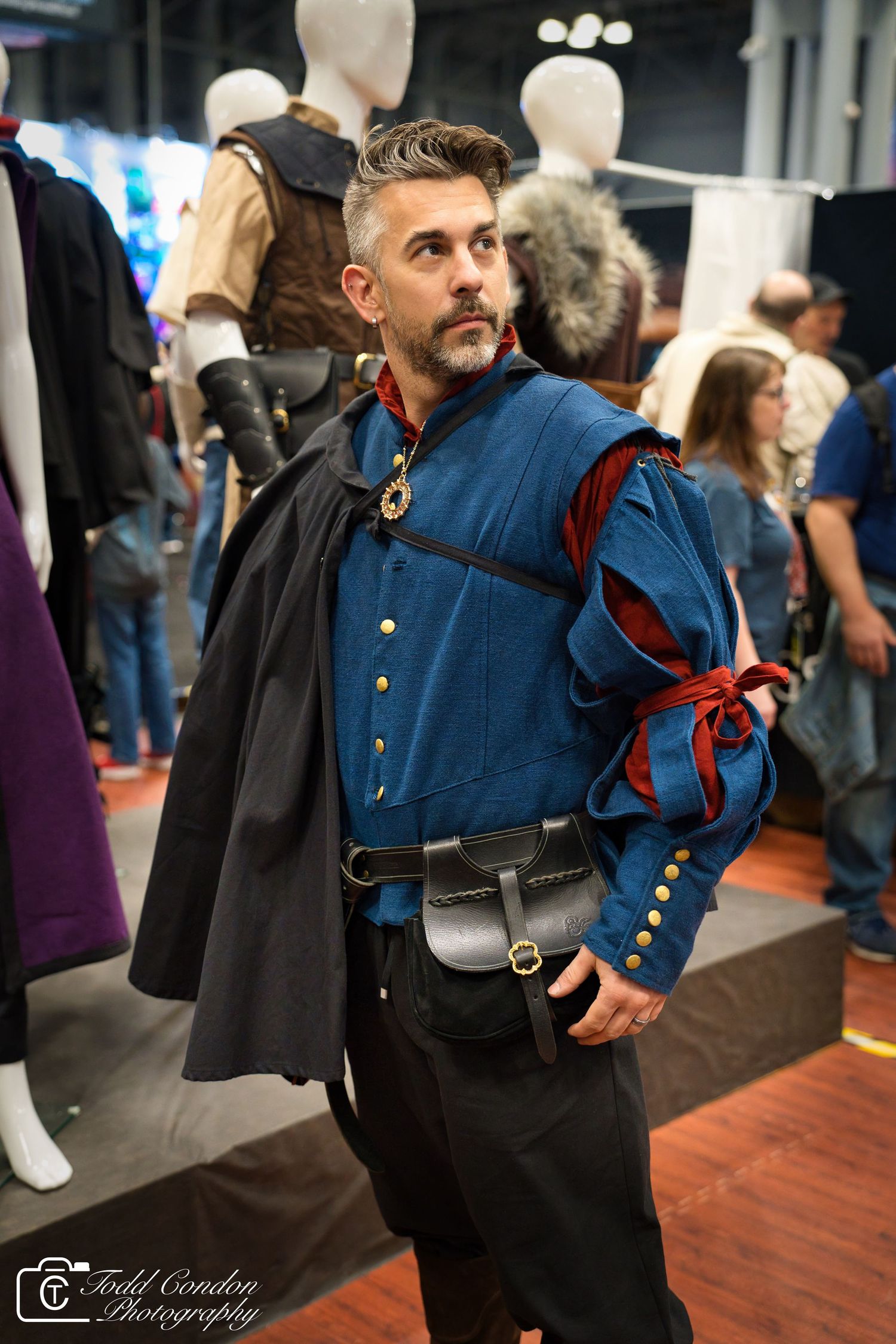 Man in Renaissance-style blue costume with cape and belt pouch at a cosplay convention.
